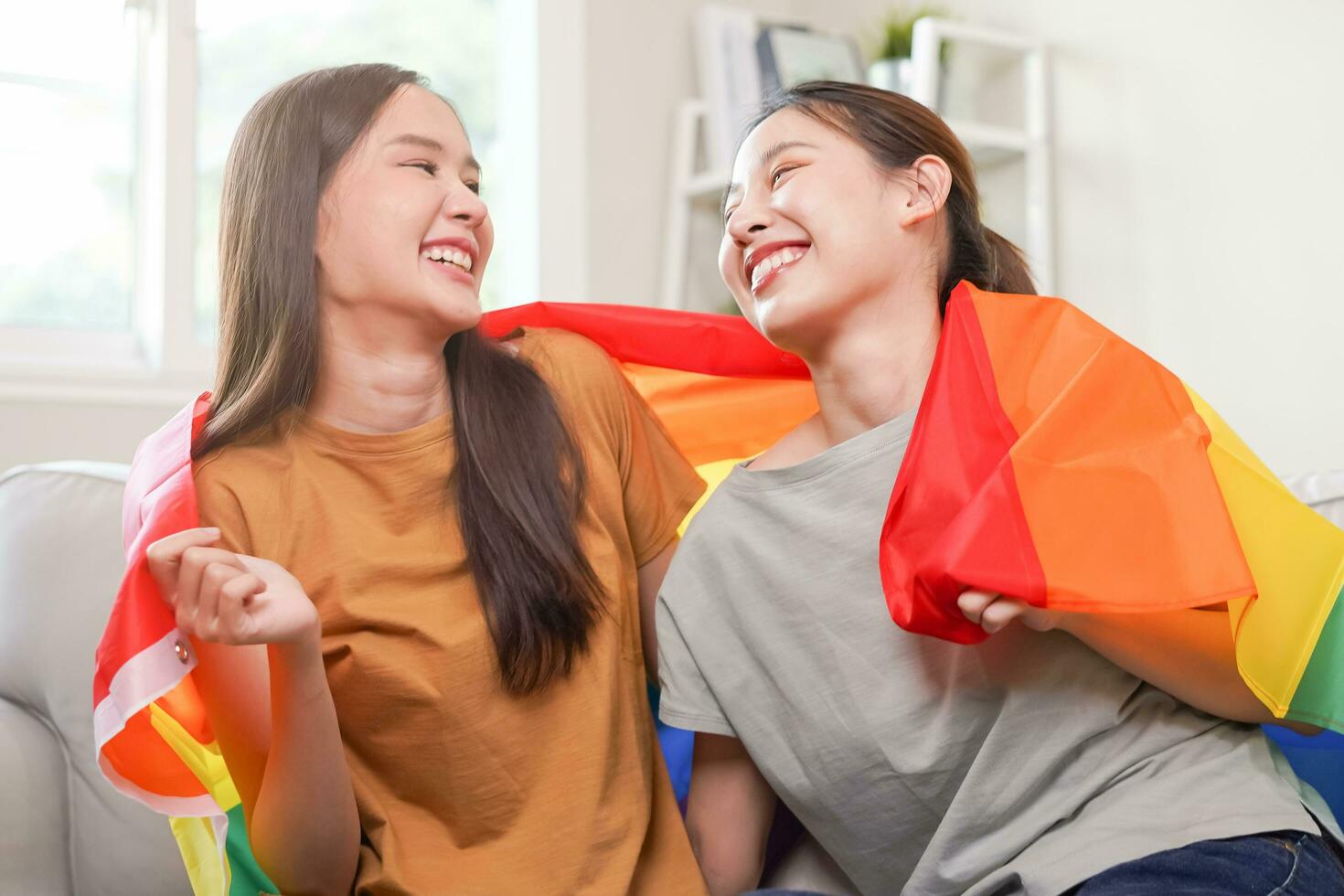 contento lesbiana, hermosa asiático joven dos mujer, niña homosexual, Pareja amor momento gasto bueno hora juntos, participación o ondulación lgbt arcoíris, orgullo bandera juntos, sentado en sofá a hogar. lgbt concepto. foto