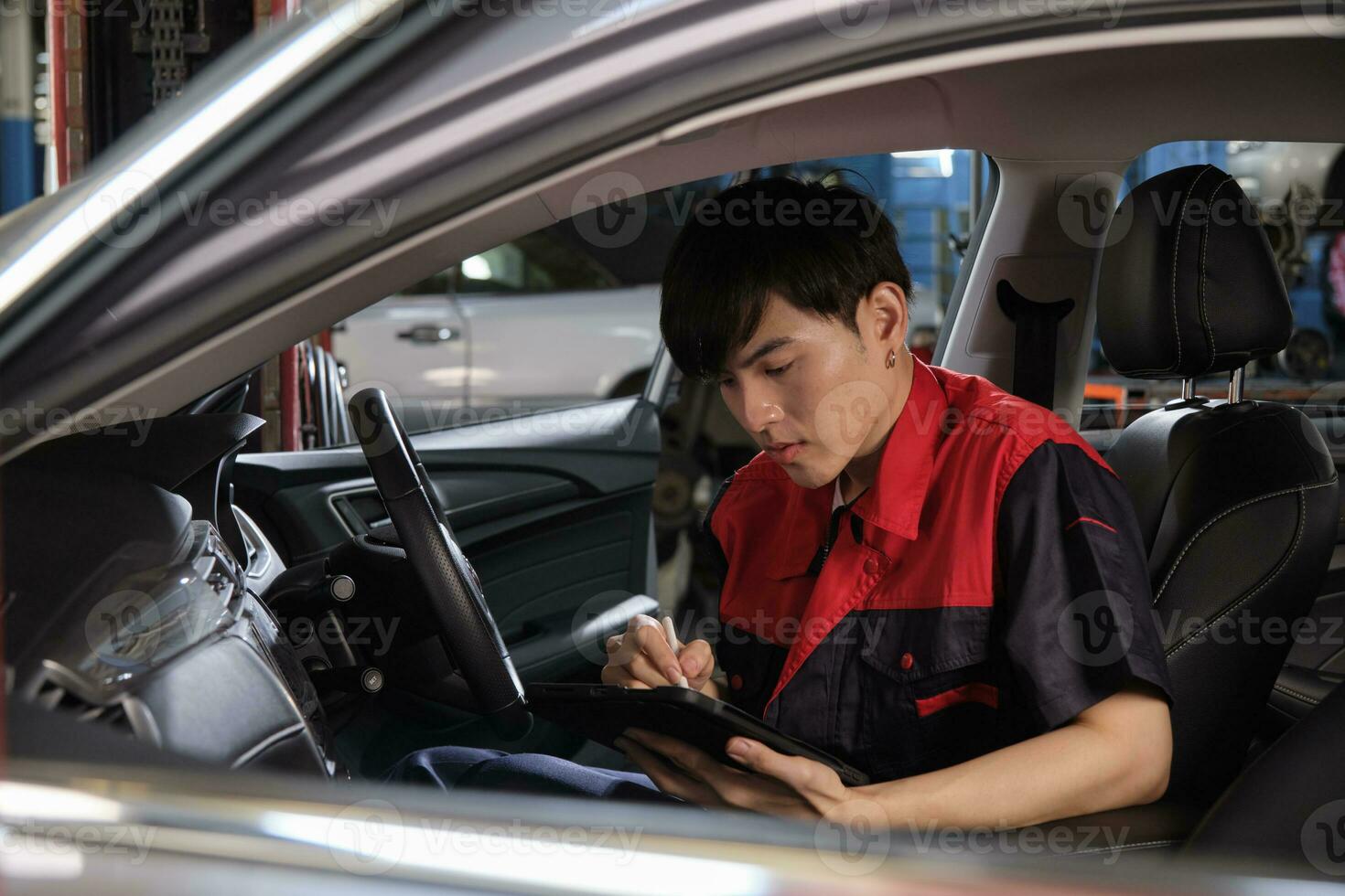 One young expert Asian male automotive mechanic technician checking a maintenance list with tablet in car interior at garage. Vehicle service fix and repair works, industrial occupation business jobs. photo