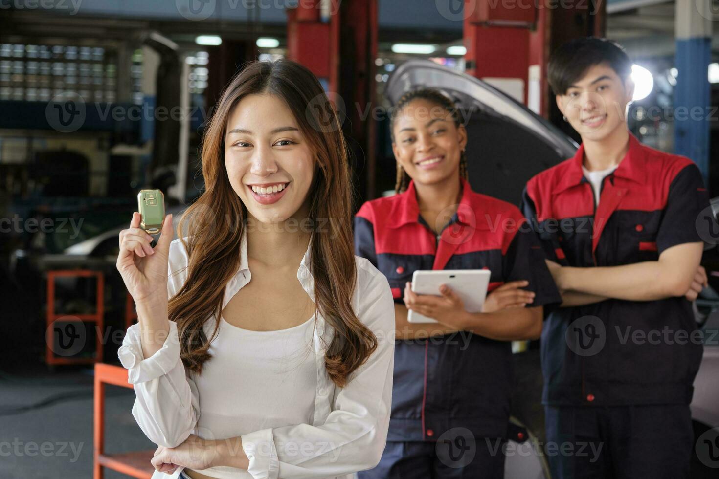 Young Asian female customer shows car key with automotive mechanic workers, happy smile in quality trust at maintenance garage, professional vehicle service center, fix check and repair auto industry. photo