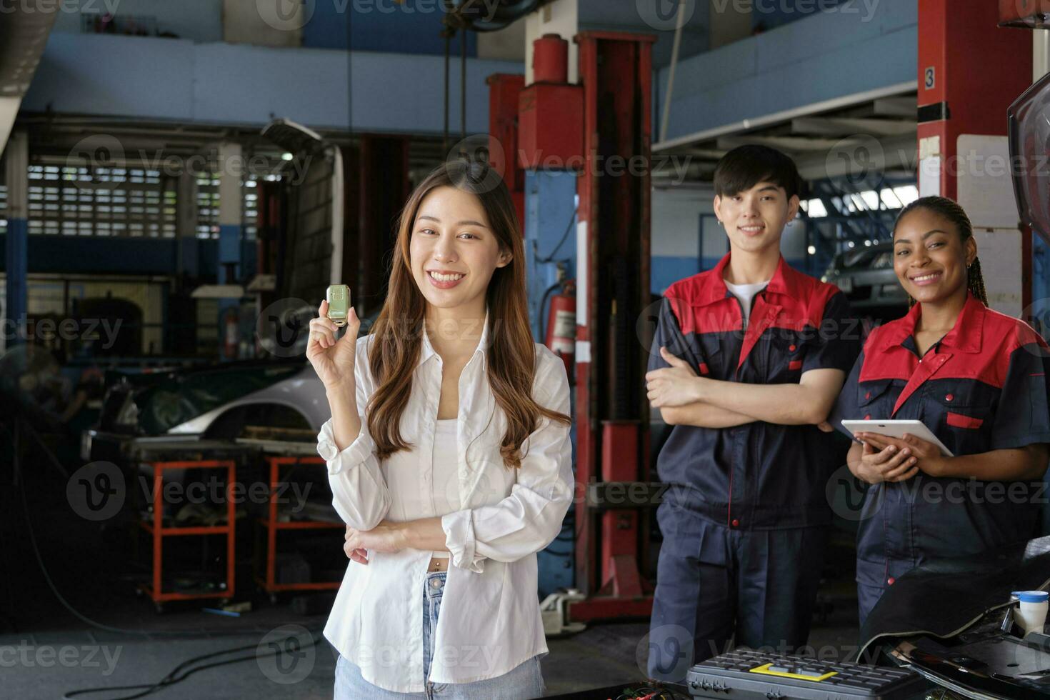 Young Asian female customer shows car key with automotive mechanic workers, happy smile in quality trust at maintenance garage, professional vehicle service center, fix check and repair auto industry. photo