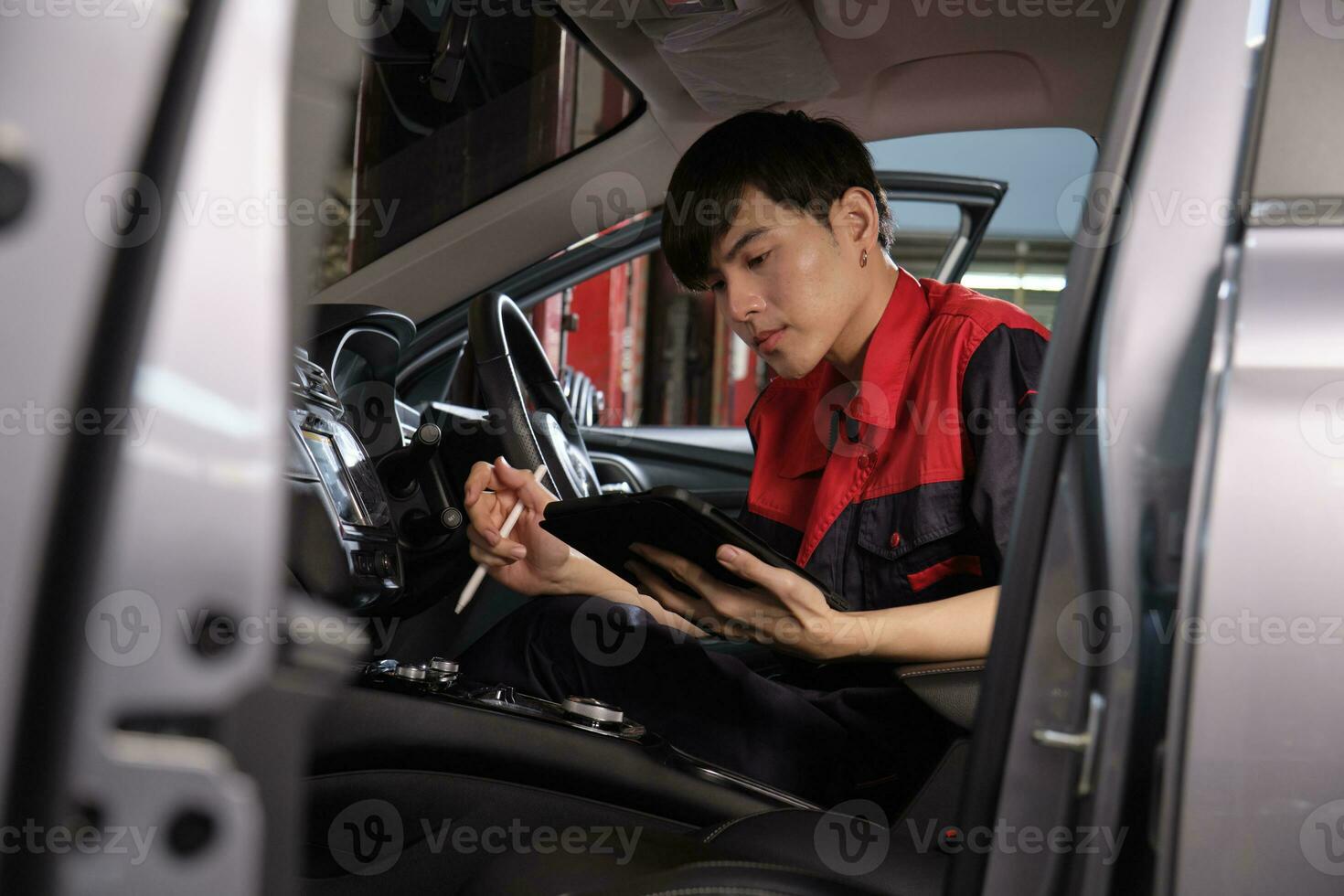 One young expert Asian male automotive mechanic technician checking a maintenance list with tablet in car interior at garage. Vehicle service fix and repair works, industrial occupation business jobs. photo