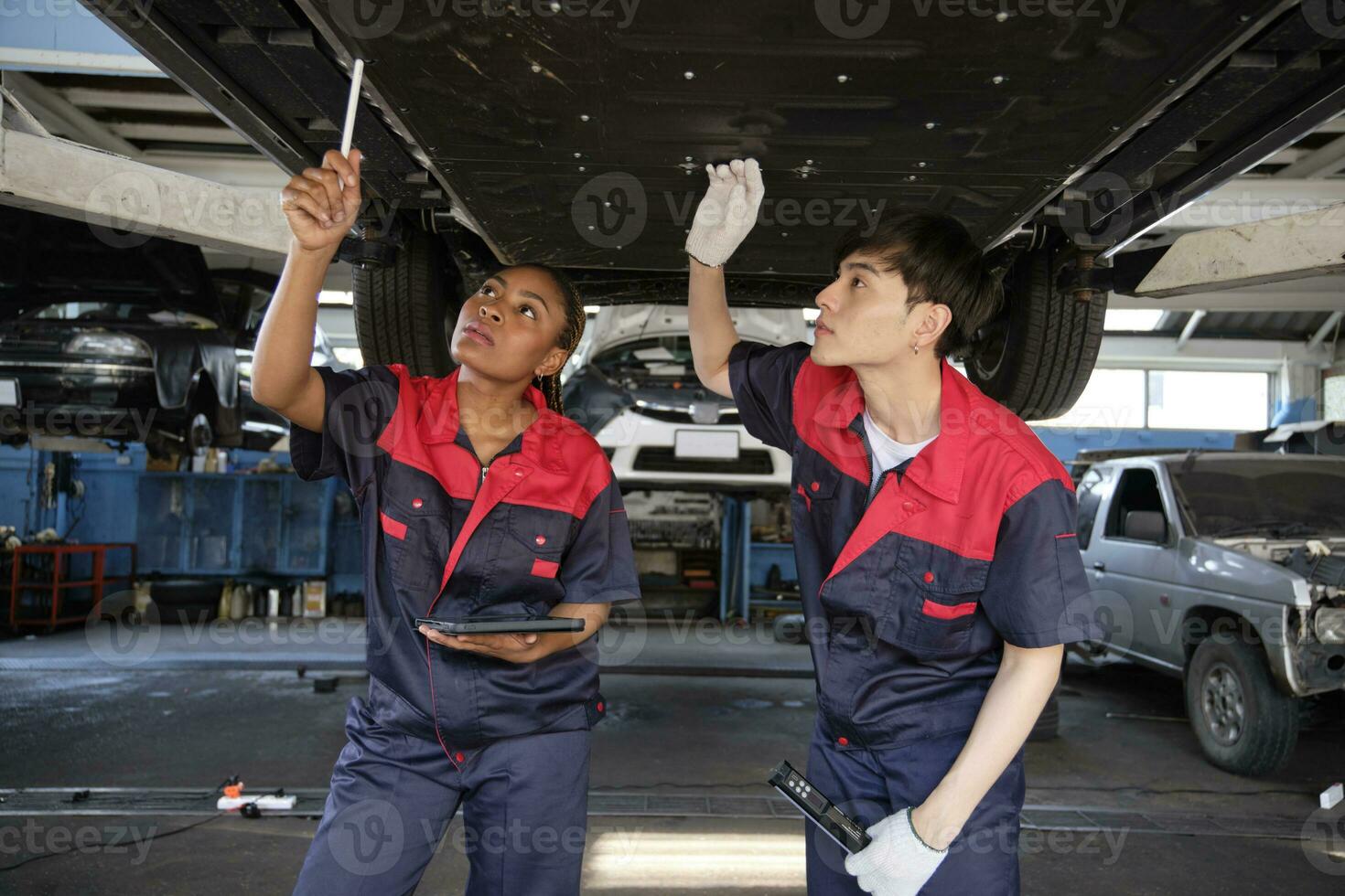Two professional automotive worker partners discuss mechanical check undercarriage of lifted electric car, fix and repair at maintenance garage, and expert service business works industry occupation. photo
