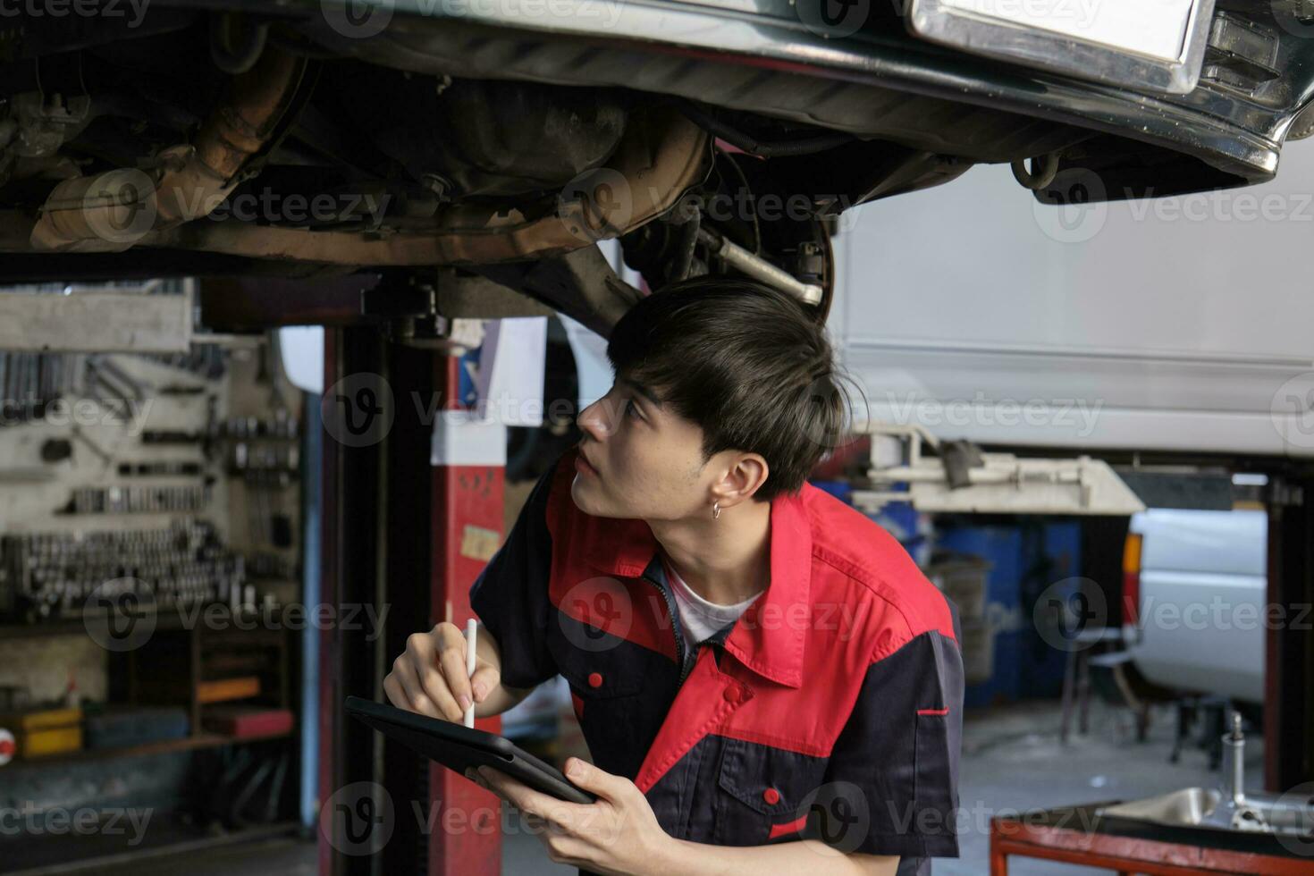 profesional joven asiático masculino motor mecánico inspecciona tren de aterrizaje de eléctrico coche levantado por máquina elevadora Jack para reparar a cochera, automotor mantenimiento Servicio trabajos industria ocupación negocio. foto