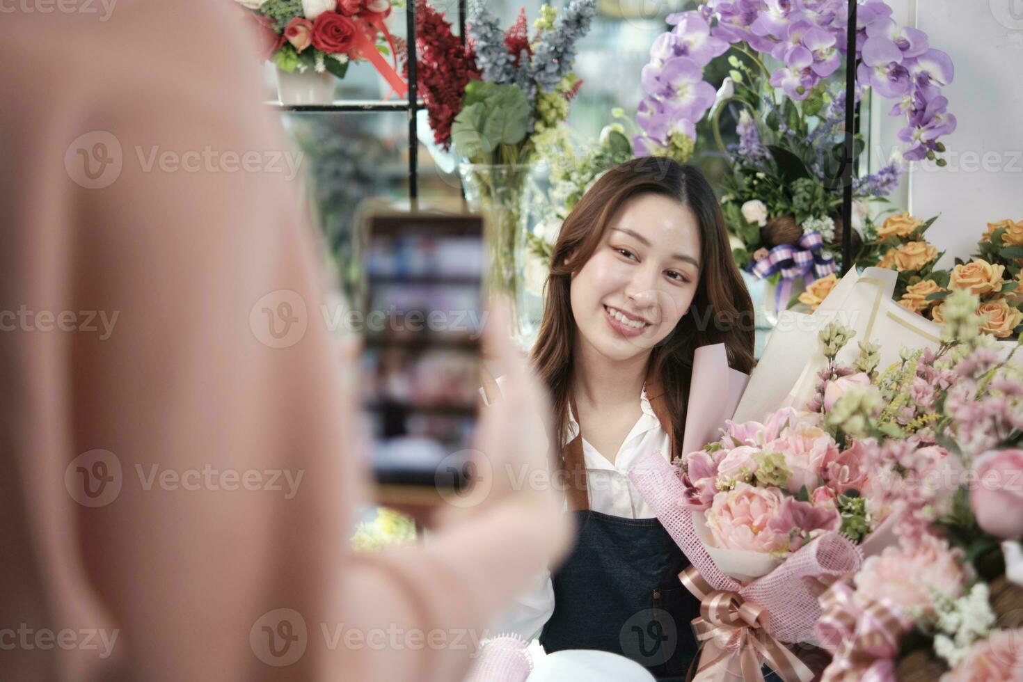 E-commerce business, young Asian female florist worker demonstrates and shows floral arrangements via online live streaming by smartphone application in bright flower shop, beautiful blossoms store. photo
