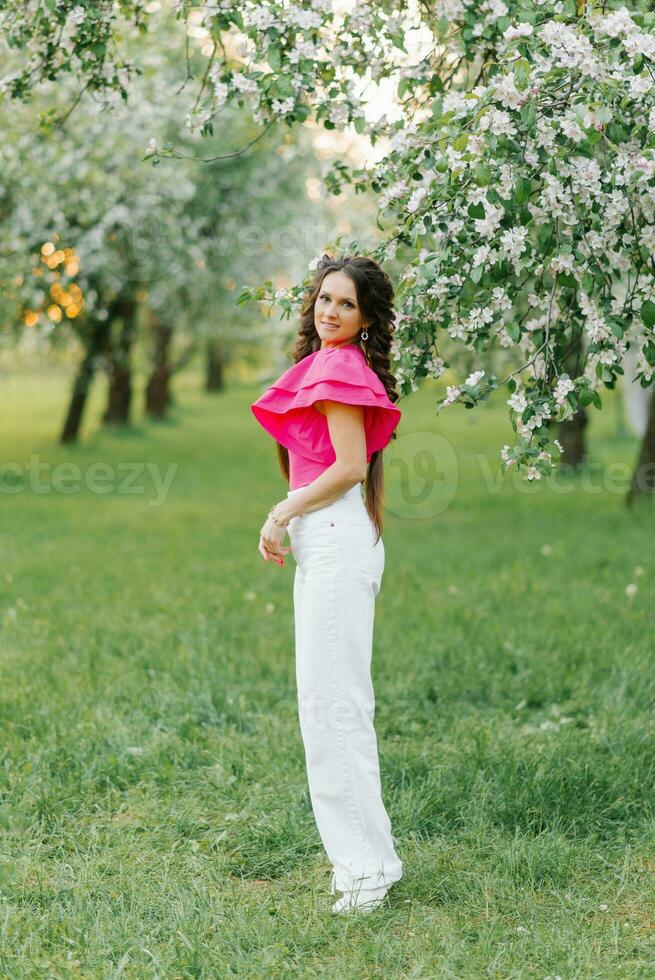 A cute young woman in white trousers and a bright pink blouse strolls through a spring park or garden among the flowering trees of an apple tree photo