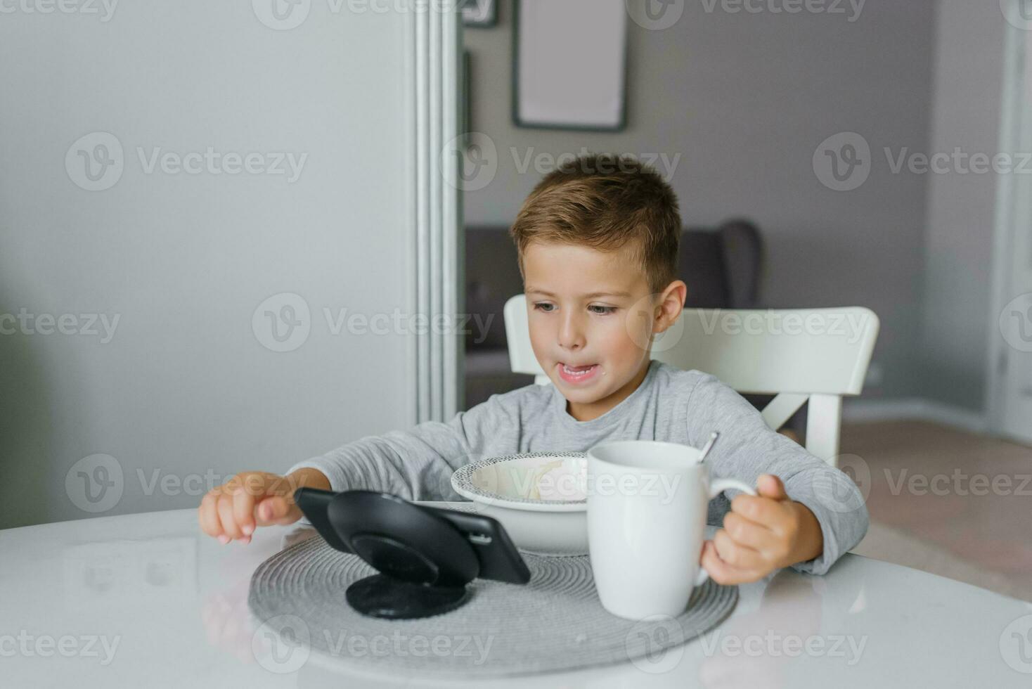 Child, a boy of five years old, is having breakfast at the kitchen table and watching cartoons on his smartphone photo