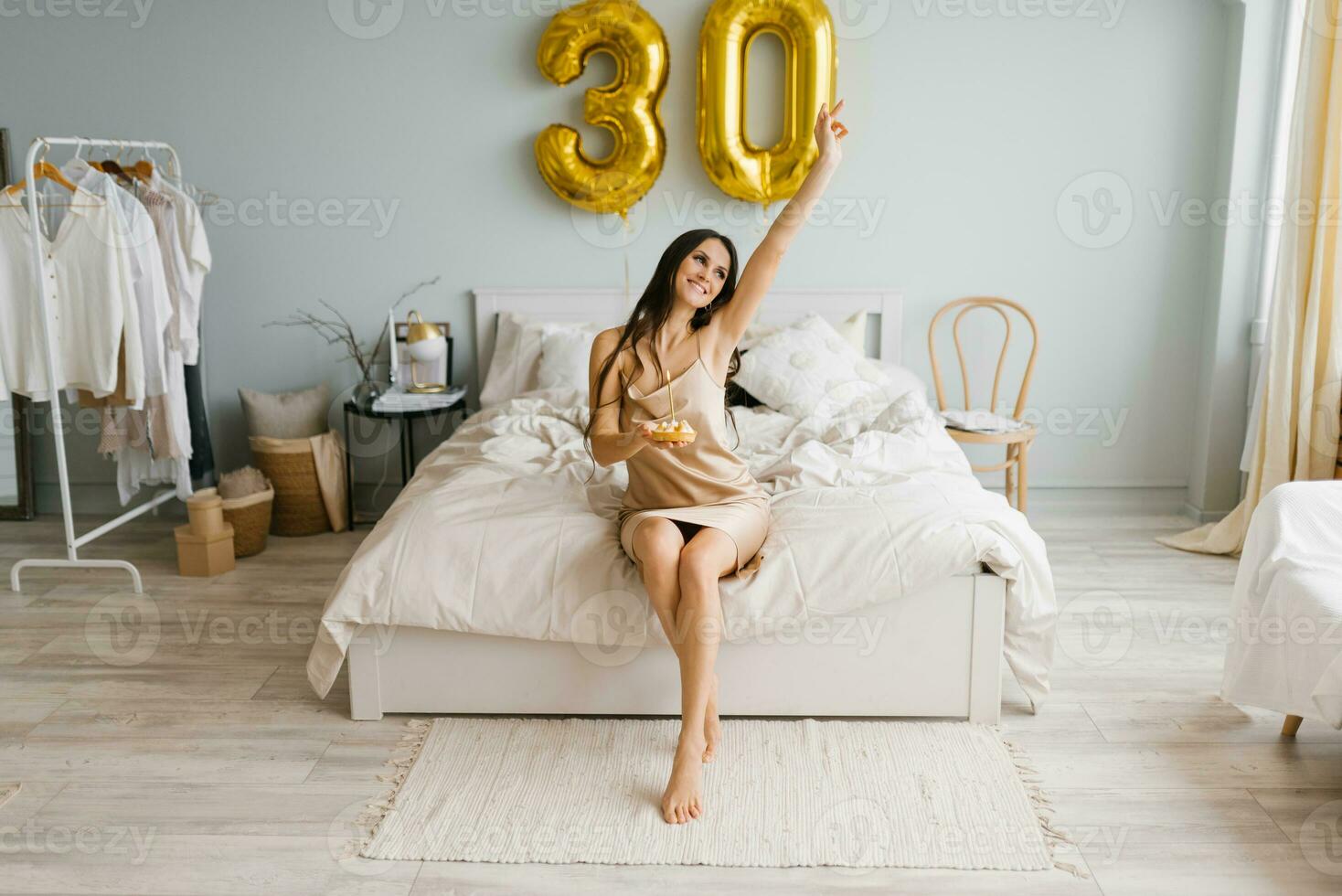 Cute woman with long hair is sitting on a bed in a cozy bedroom at home with a cake on a plate and a candle and celebrating her birthday photo
