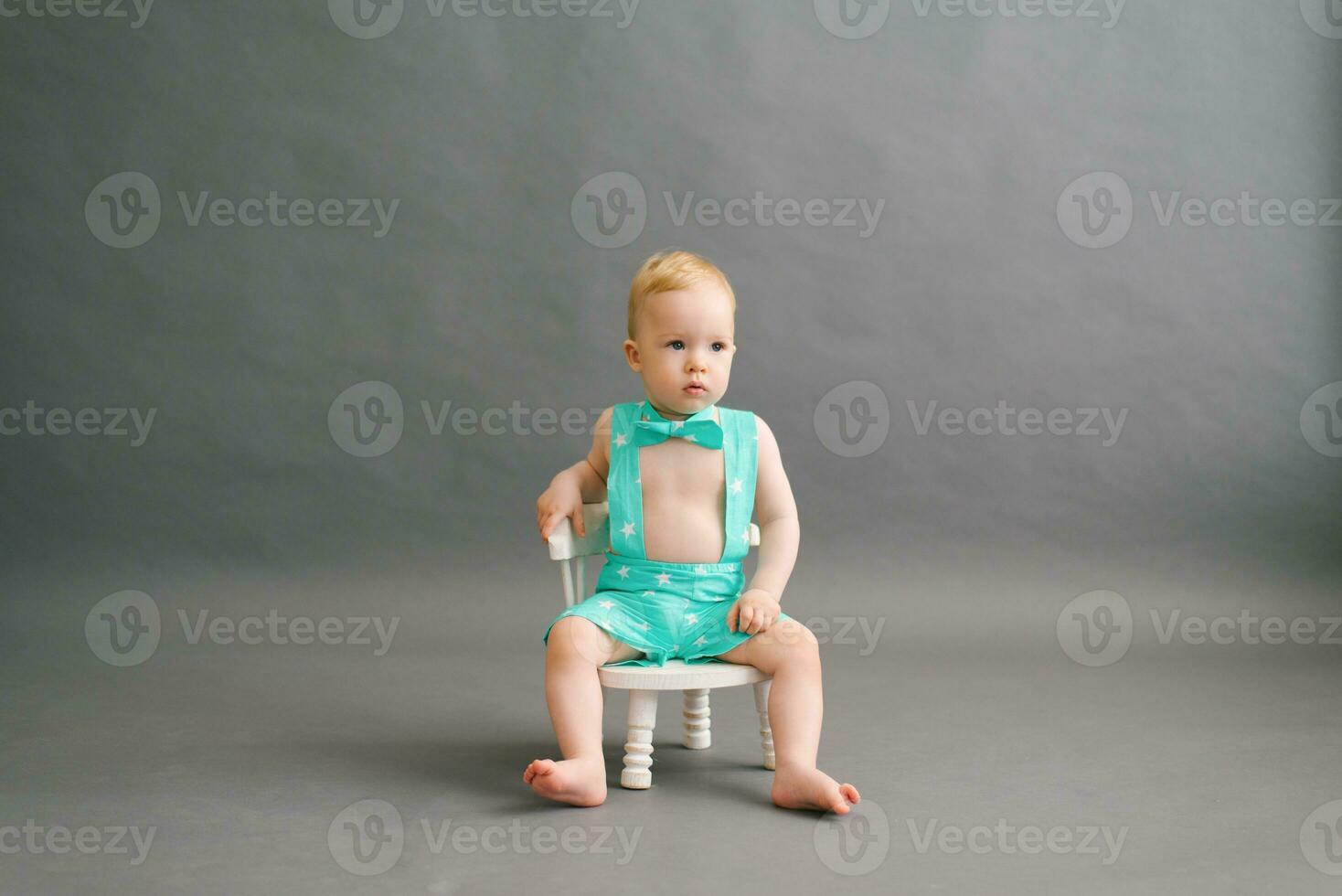 A funny little boy in a summer suit is sitting on a white children's chair on a gray background in a photo studio