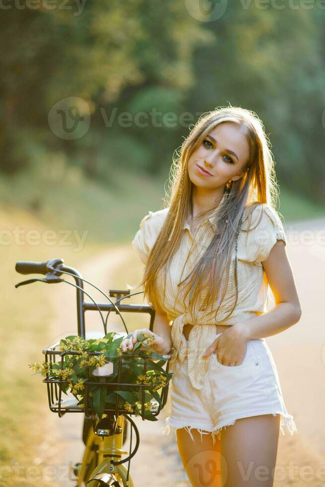 Young beautiful woman standing next to a bicycle with a wicker basket full of flowers in the forest photo