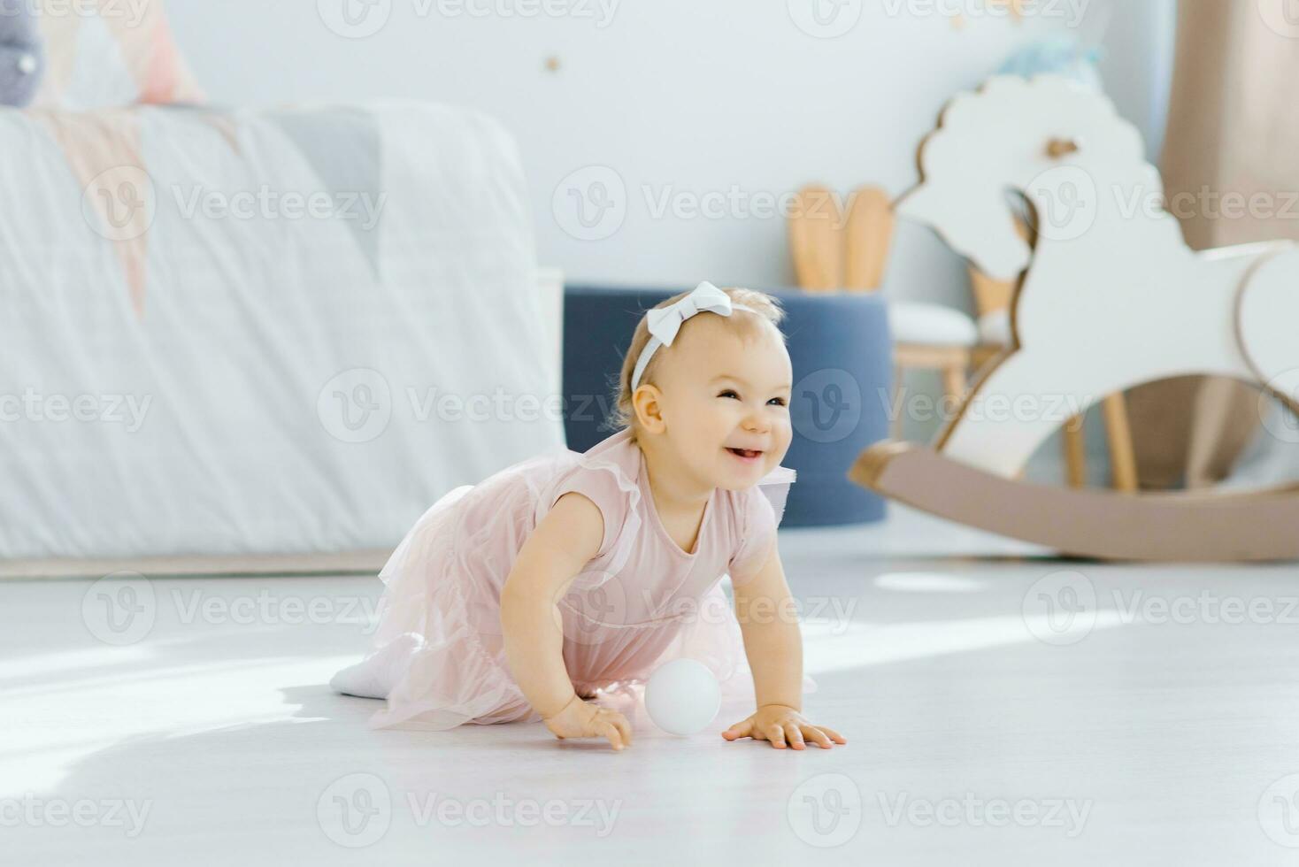 A pretty blue eyed blonde one year old in a pink dress crawls on all fours in the nursery on the floor and smiles fervently photo