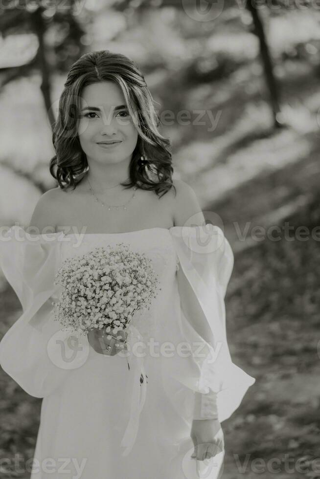 increíble novia en un largo Boda vestir con un hermosa ramo de flores en mano y con encantador sonrisa. debajo el árbol en el hermosa puesta de sol ligero en el parque. foto