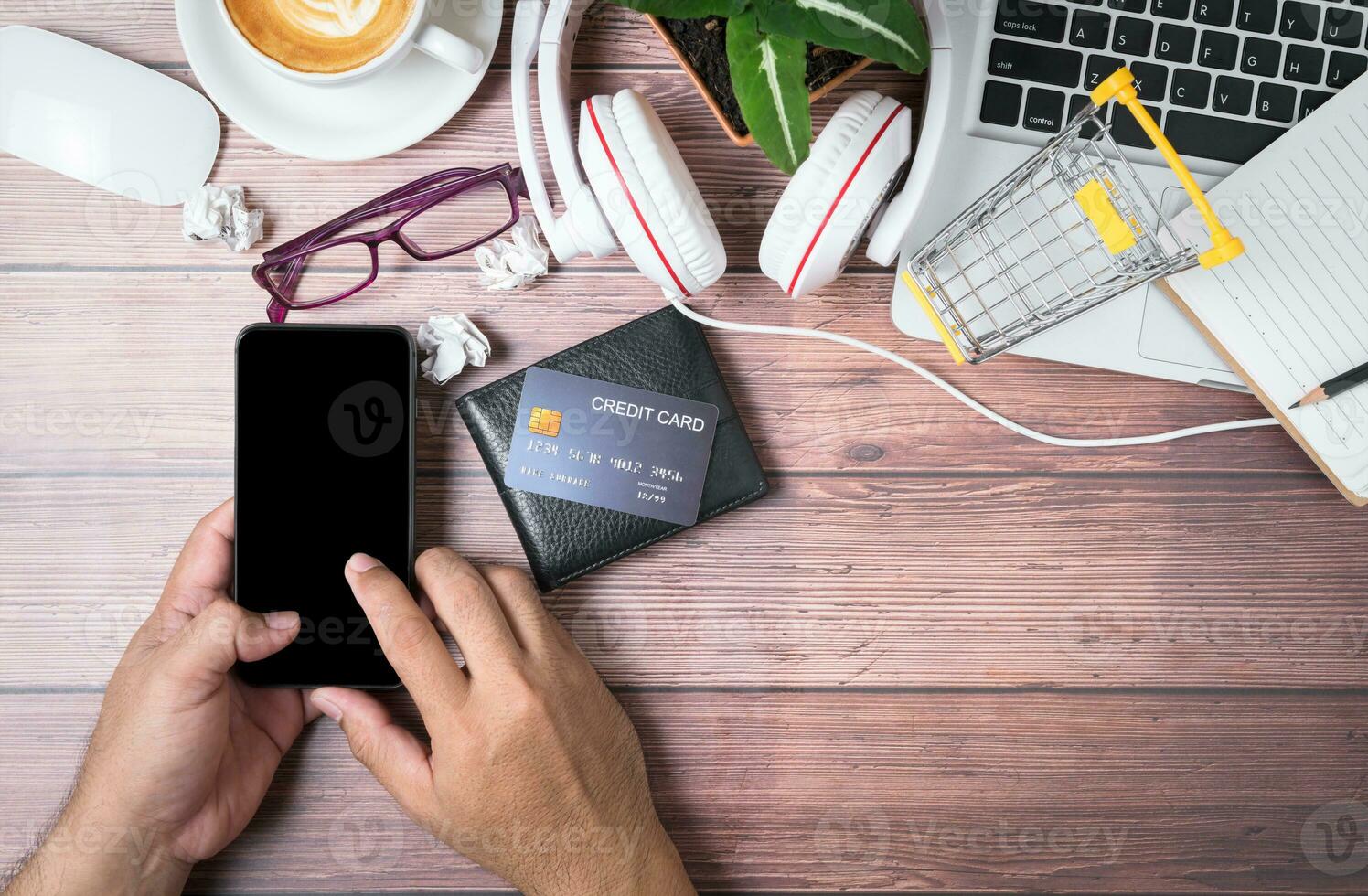 man touch on black screen mobile phone with credit card on wallet and laptop on wooden photo