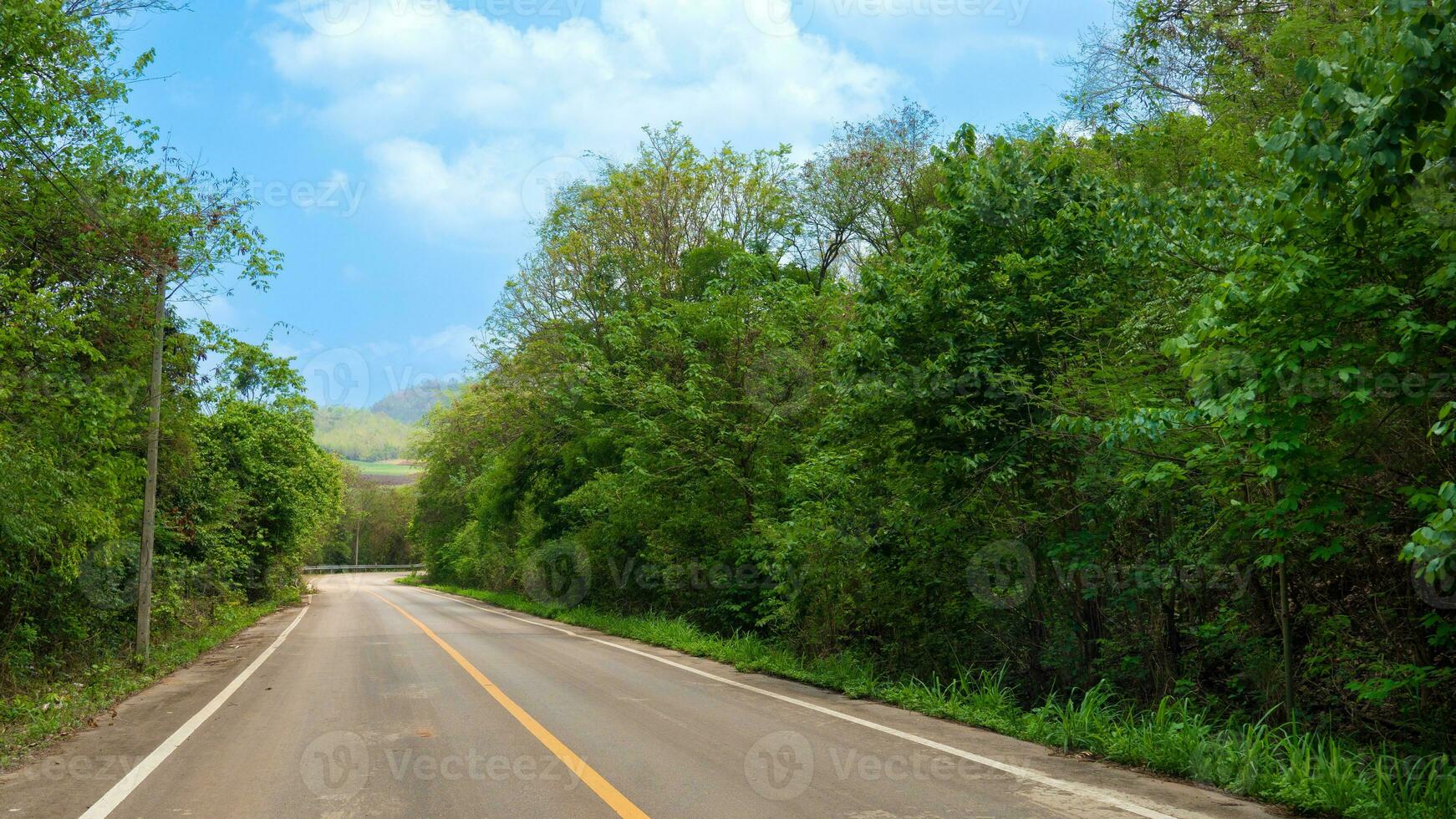 Road goes straight ahead of the asphalt road and the curve at the end in Thailand. Two besie with green forest and under blue sky and white clouds. photo