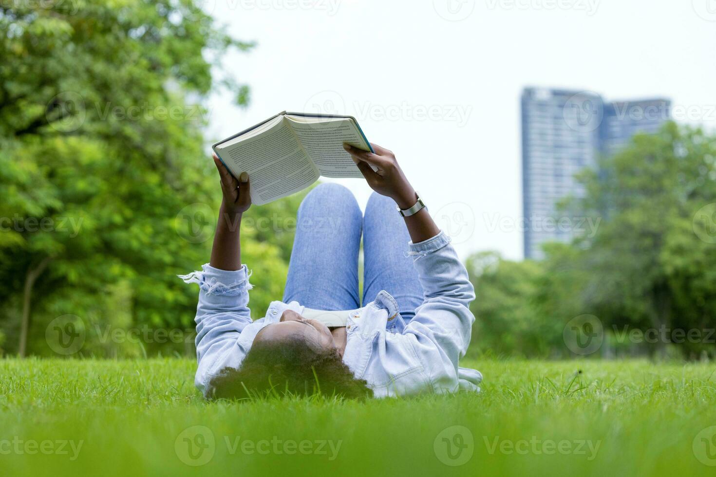 African American woman is lying down in the grass lawn inside the public park holding book in her hand during summer for reading and education concept photo