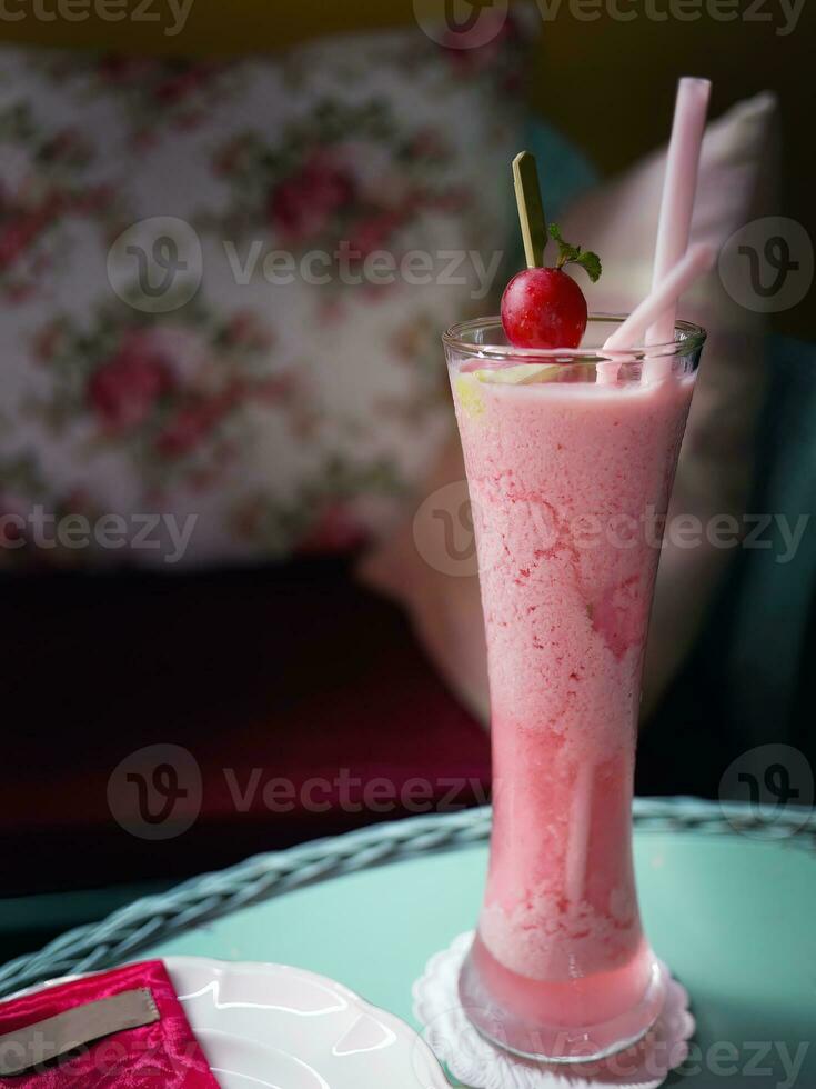 Cold red fruit juice in a tall glass decorated with 2 straws and a cherry on a blue table, selective focus with dark background, flowers cafe concept photo