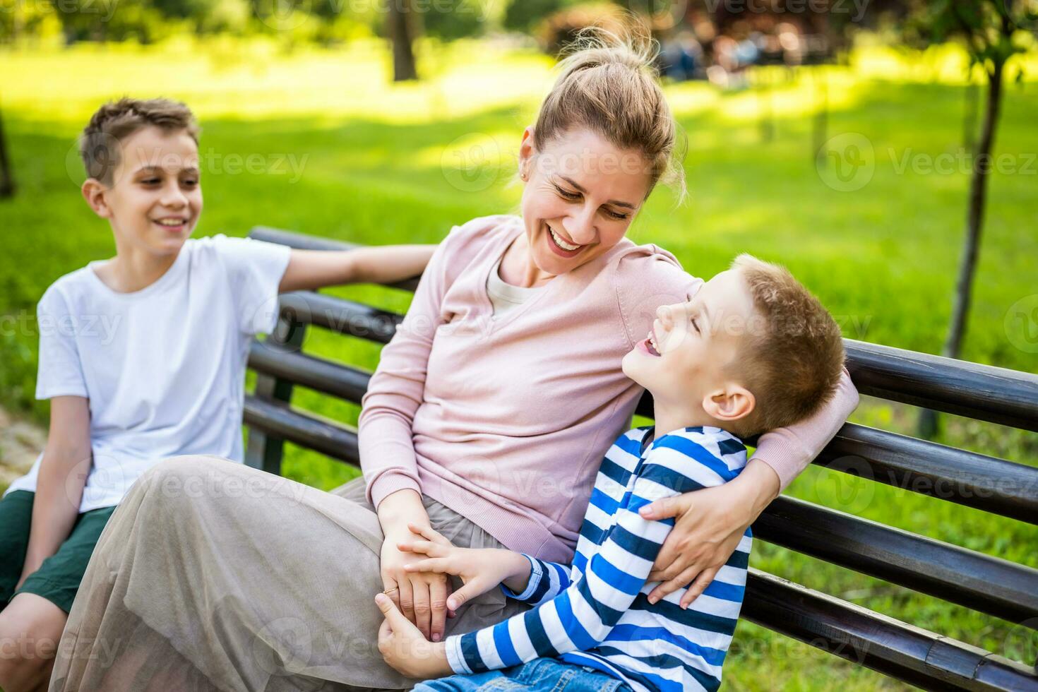 Happy mother is sitting with her sons on bench in park. They are having fun together. photo