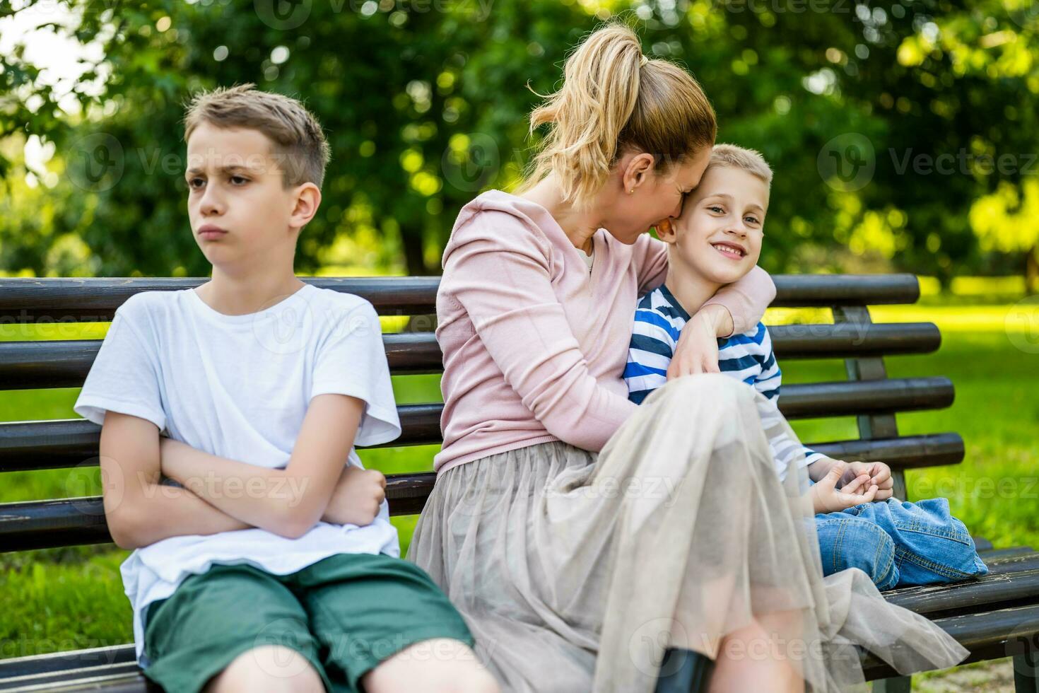 Happy mother is sitting with her sons on bench in park. One boy is offended and pouting. photo