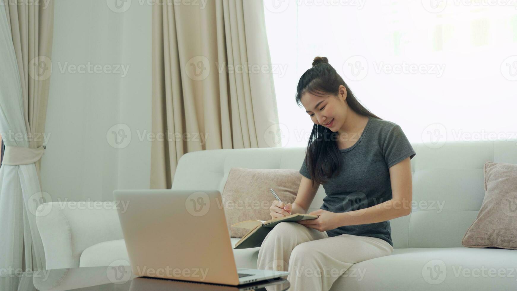Asian woman sits on the sofa while taking notes from her computer screen while working or studying online at home. photo