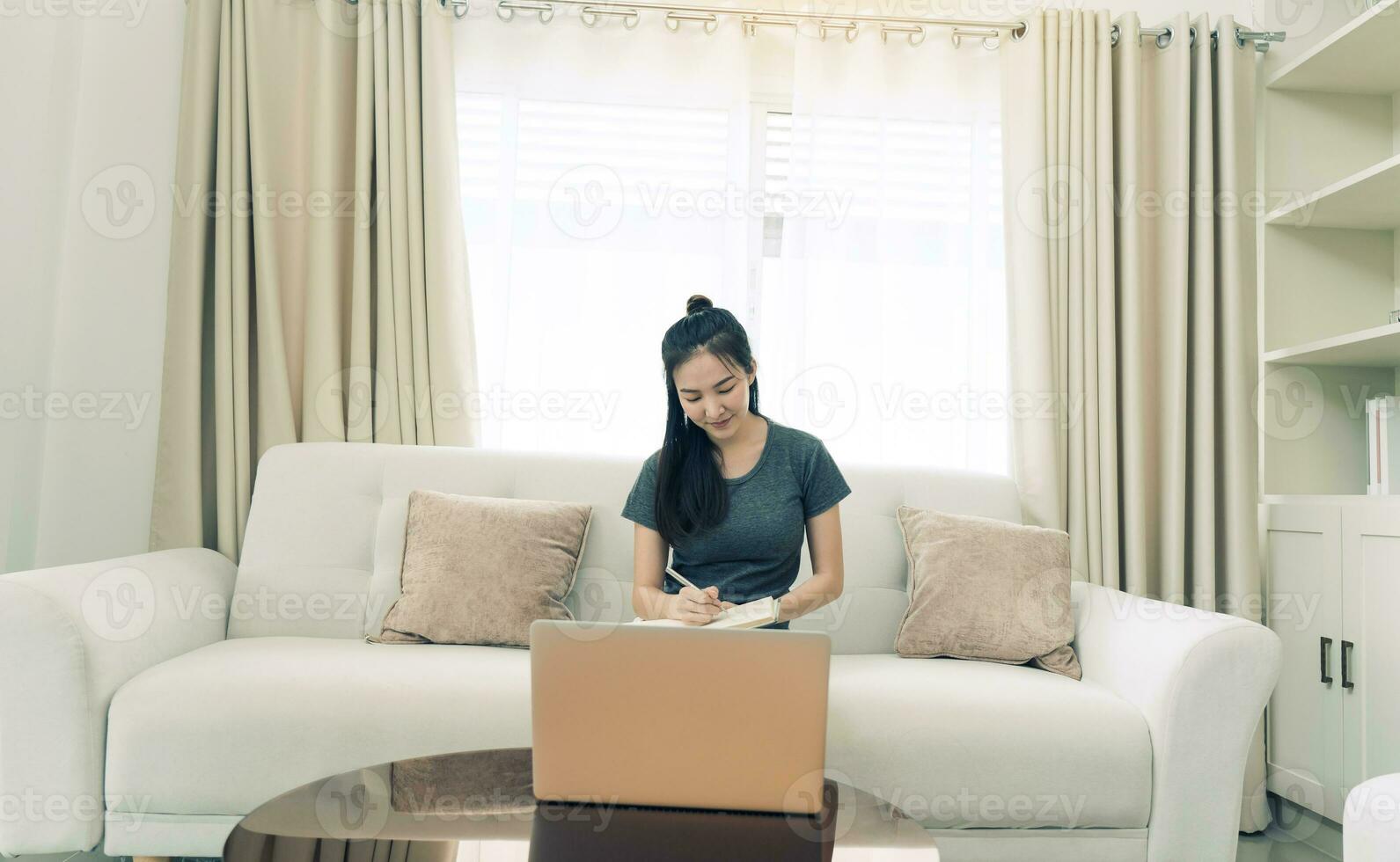 una mujer asiática se sienta en el sofá mientras toma notas de la pantalla de su computadora mientras trabaja o estudia en línea en casa. foto