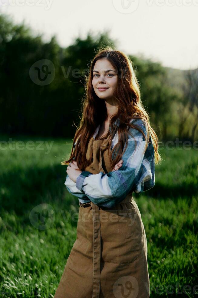 un granjero mujer en su trabajo ropa, tartán camisa y delantal, soportes en el campo en el verde césped y sonrisas en el ajuste Dom foto