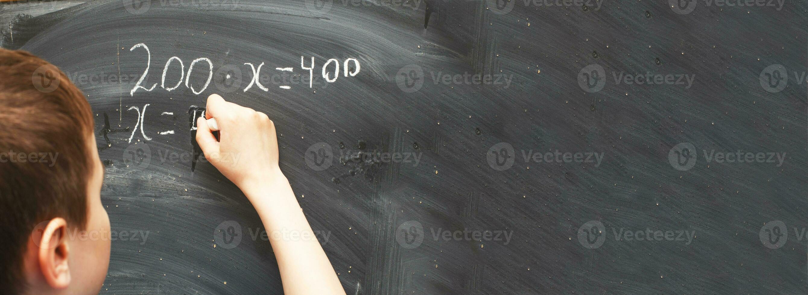 Boy standing back in front of school blackboard and writing. Schoolboy solves math example at the chalkkboard photo