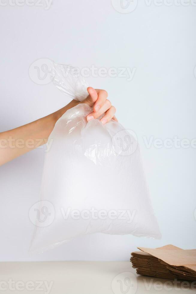 Hand holding an empty transparent plastic bag and a stack of eco-friendly paper bags on the table photo