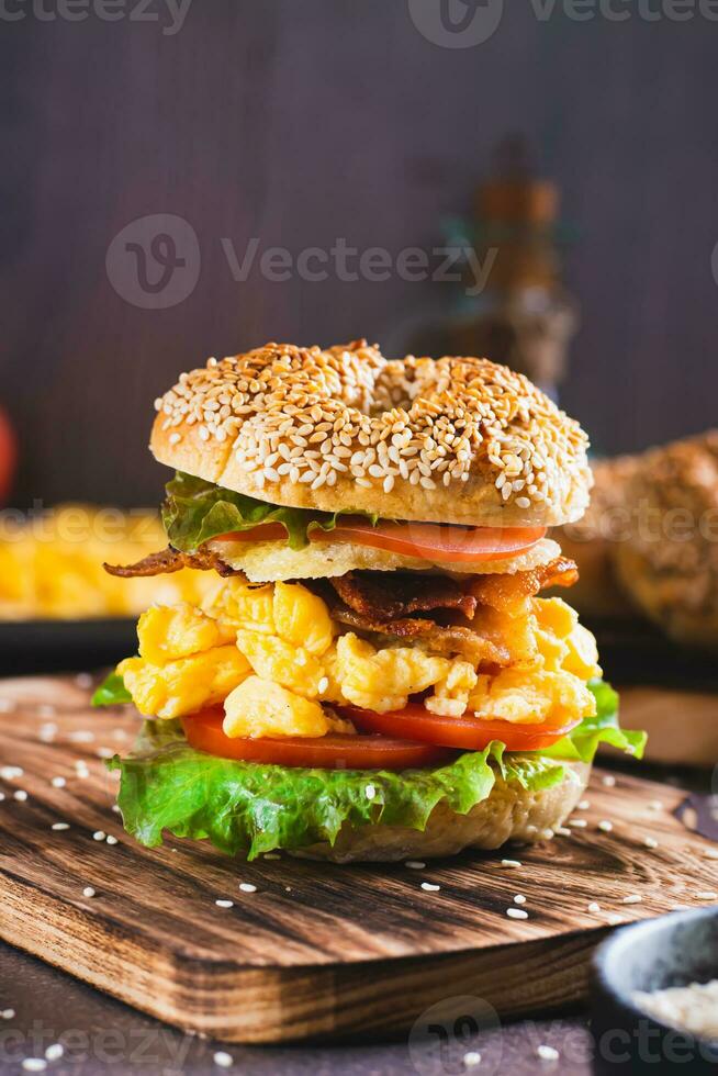 Homemade bagel with scramble egg, bacon, cheese, tomato and lettuce on a board vertical view photo