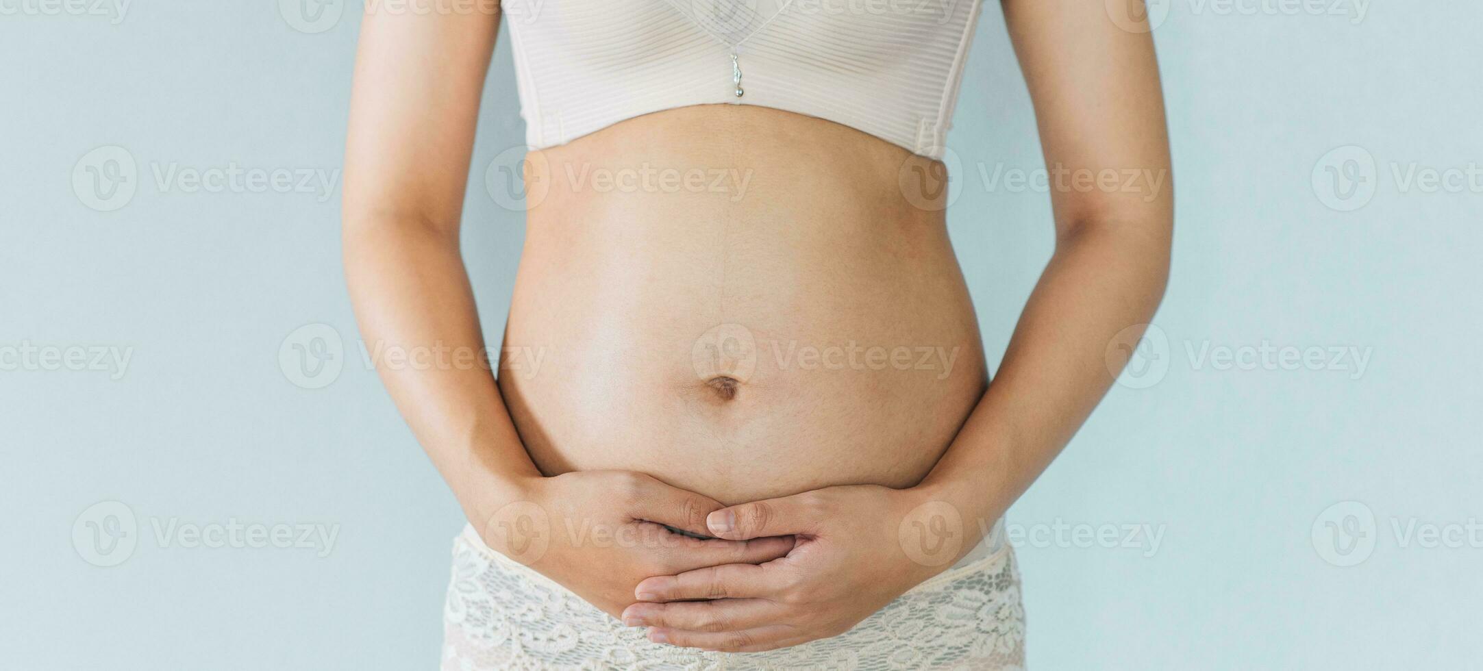 Pregnant woman holds hands on belly and pastel color background. Pregnancy, maternity, preparation and expectation concept. Close-up, copy space. Young woman waiting for baby birth. photo