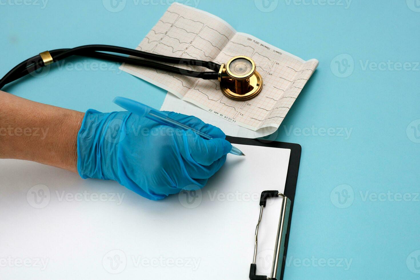 a doctor's hand in a glove writes something on a tablet with a paper with a pen next to it lies a phonendoscope and a cardiogram photo
