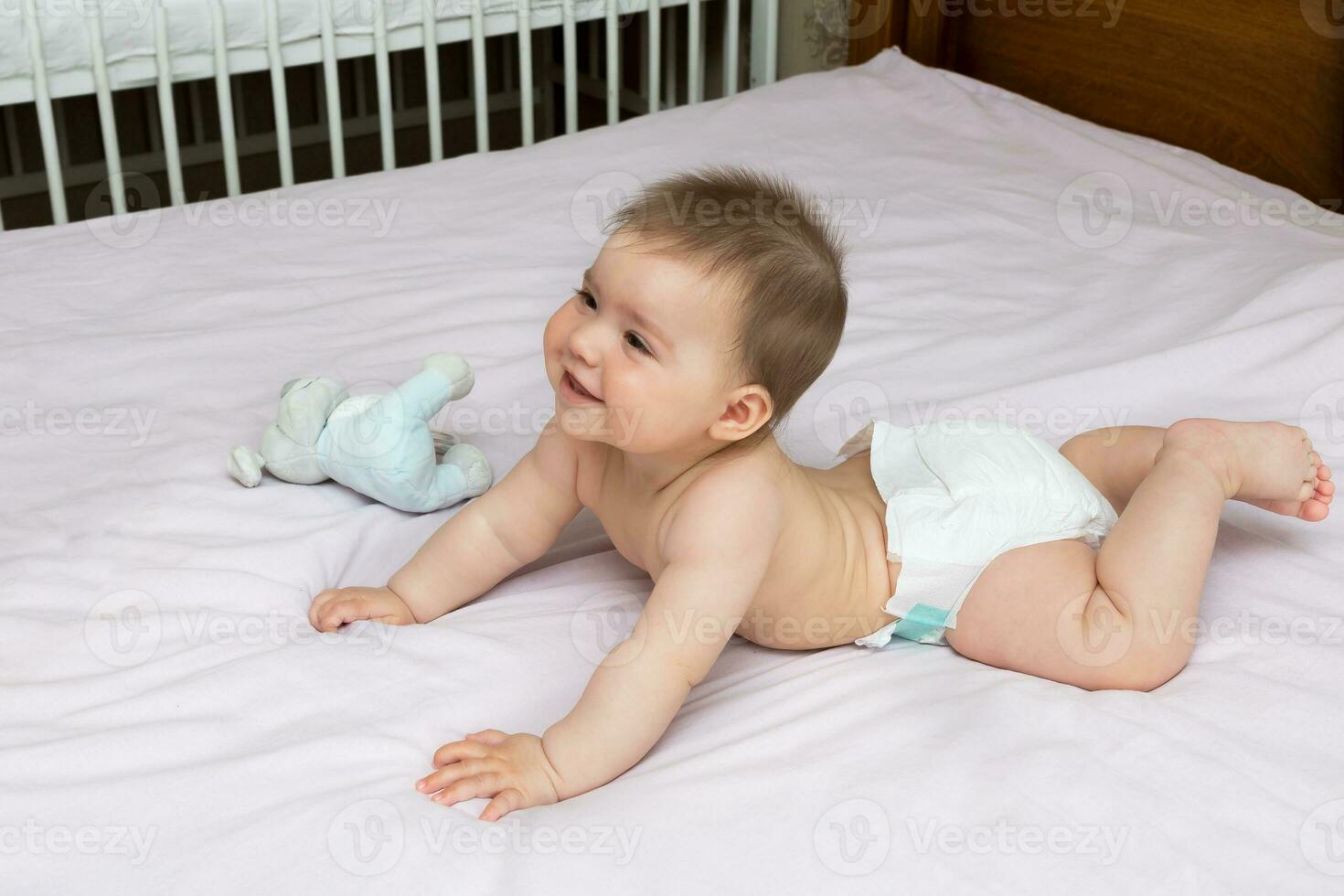 bebé niña en un pañal mentiras en su barriguita en cama con un juguete y sonrisas foto