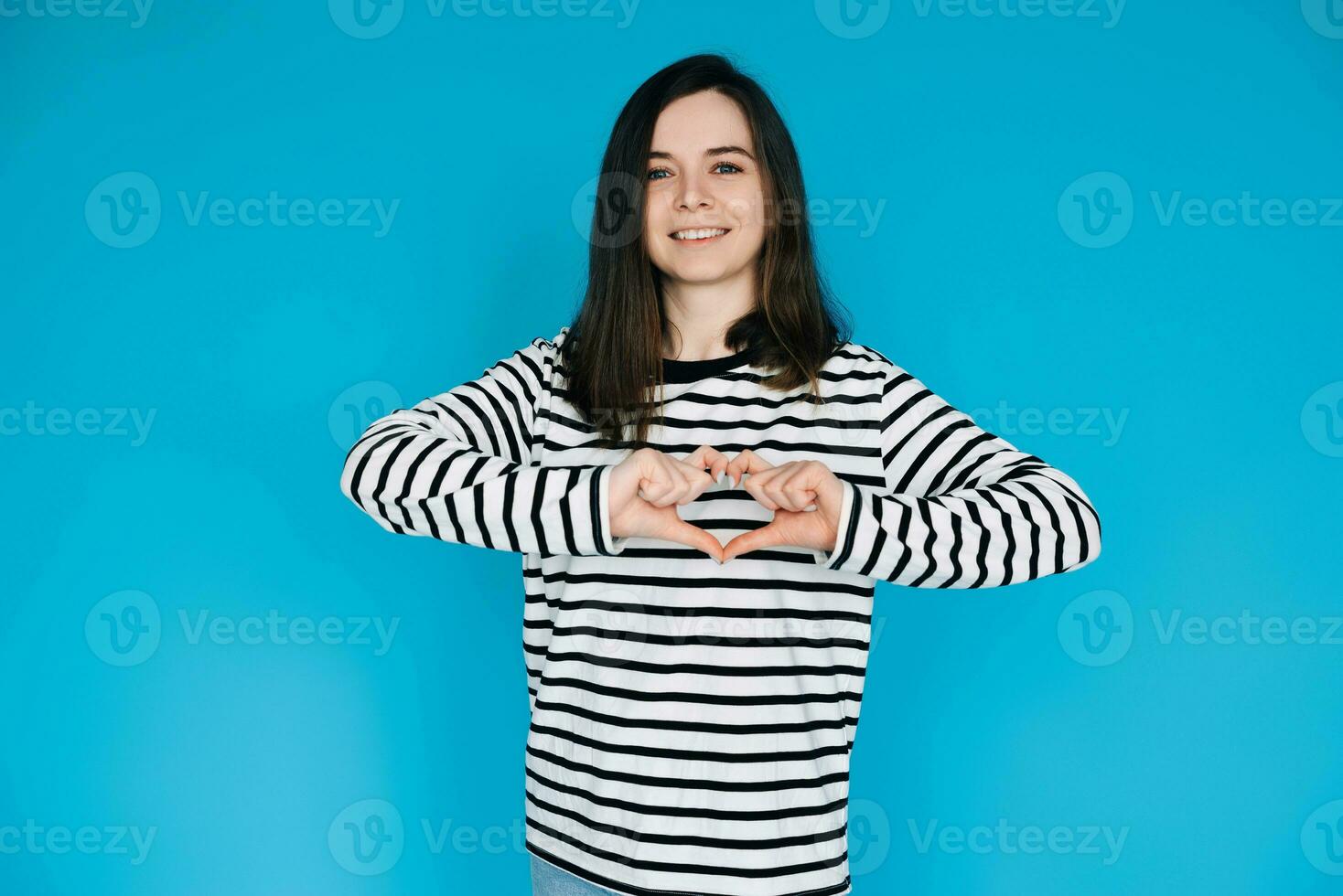 Joyful Woman in Striped Sweater Smiling, Gesturing Heart Hands - Symbol of Love and Happiness - Isolated on Blue Background - Perfect for Positive Emotions, Love, and Lifestyle Concepts photo