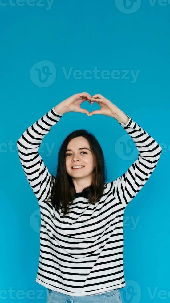 Joyful Woman in Striped Sweater Expressing Love and Happiness - Happy Girl Showing Heart Gesture with Arms, Smiling - Isolated Blue Background with Copy Space - Perfect for Love, Happiness Concepts photo