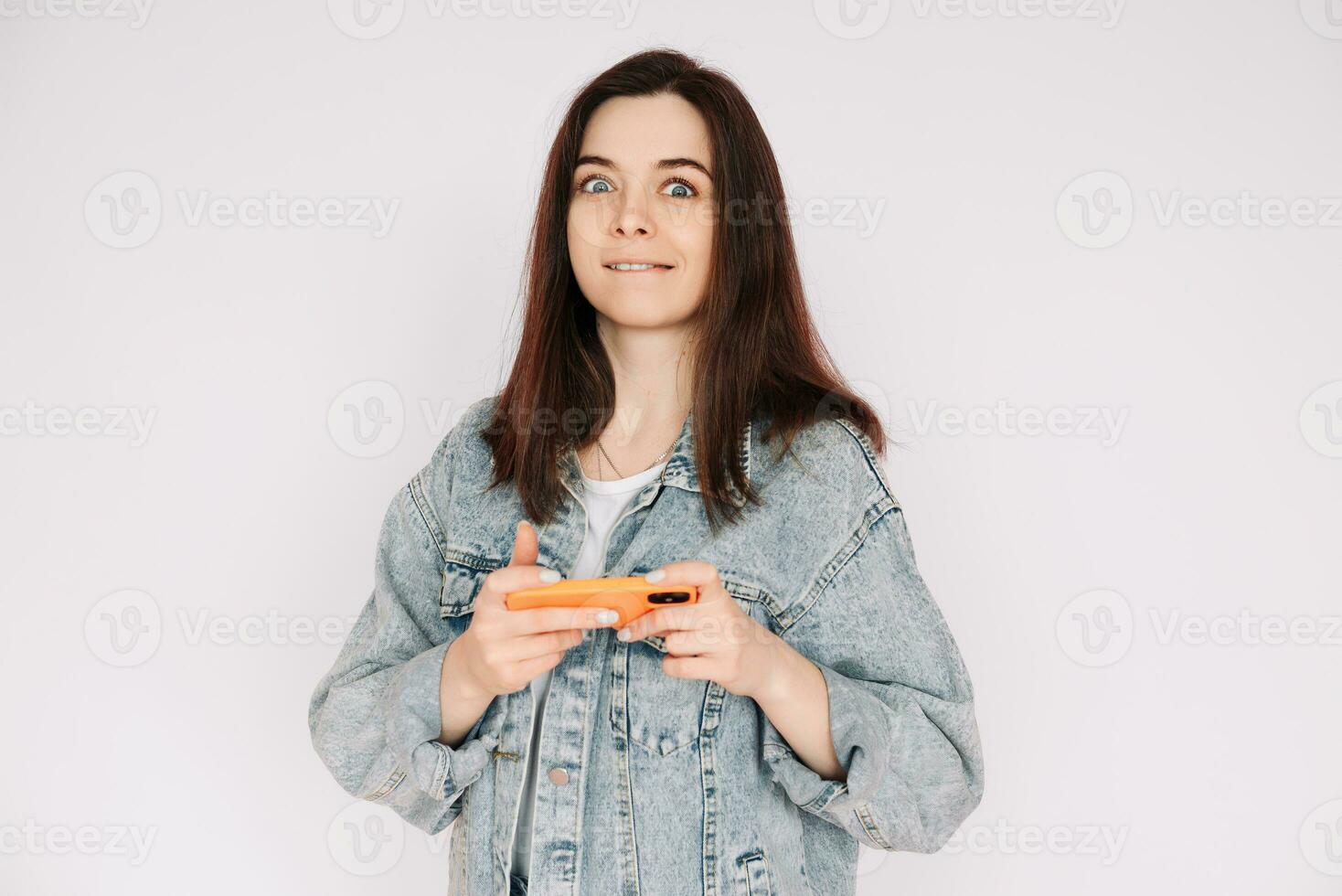 retrato de un joven mujer en un casual mezclilla chaqueta con un teléfono inteligente jugando un juego, aislado en contra un gris fondo, transporte indecisión y inseguridad foto