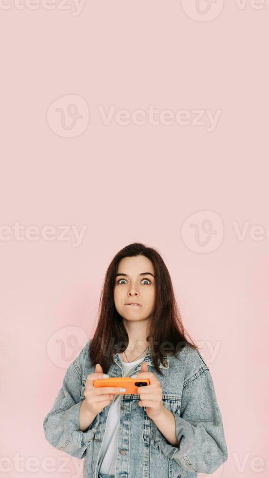 Portrait of a young woman in casual denim jacket with smartphone playing game, isolated on pink background conveying hesitancy and insecurity photo