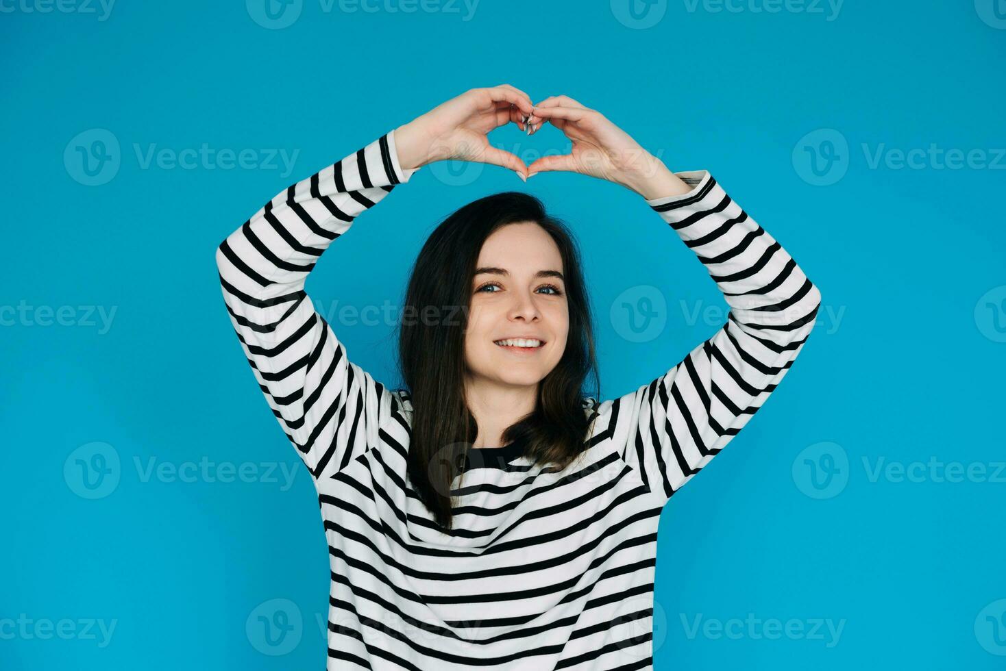 Joyful Woman in Striped Sweater Spreading Love - Smiling Girl with Raised Arms Creating Heart Shape, Radiating Happiness and Positivity - Isolated Blue Background - Perfect for Positive Vibes Concepts photo