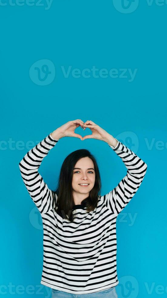 Happy Woman in Striped Sweater Spreading Love and Positivity - Smiling Girl with Raised Arms Forming Heart Shape, Expressing Joy and Affection - Isolated Blue Background - Ideal for Happiness, Love photo