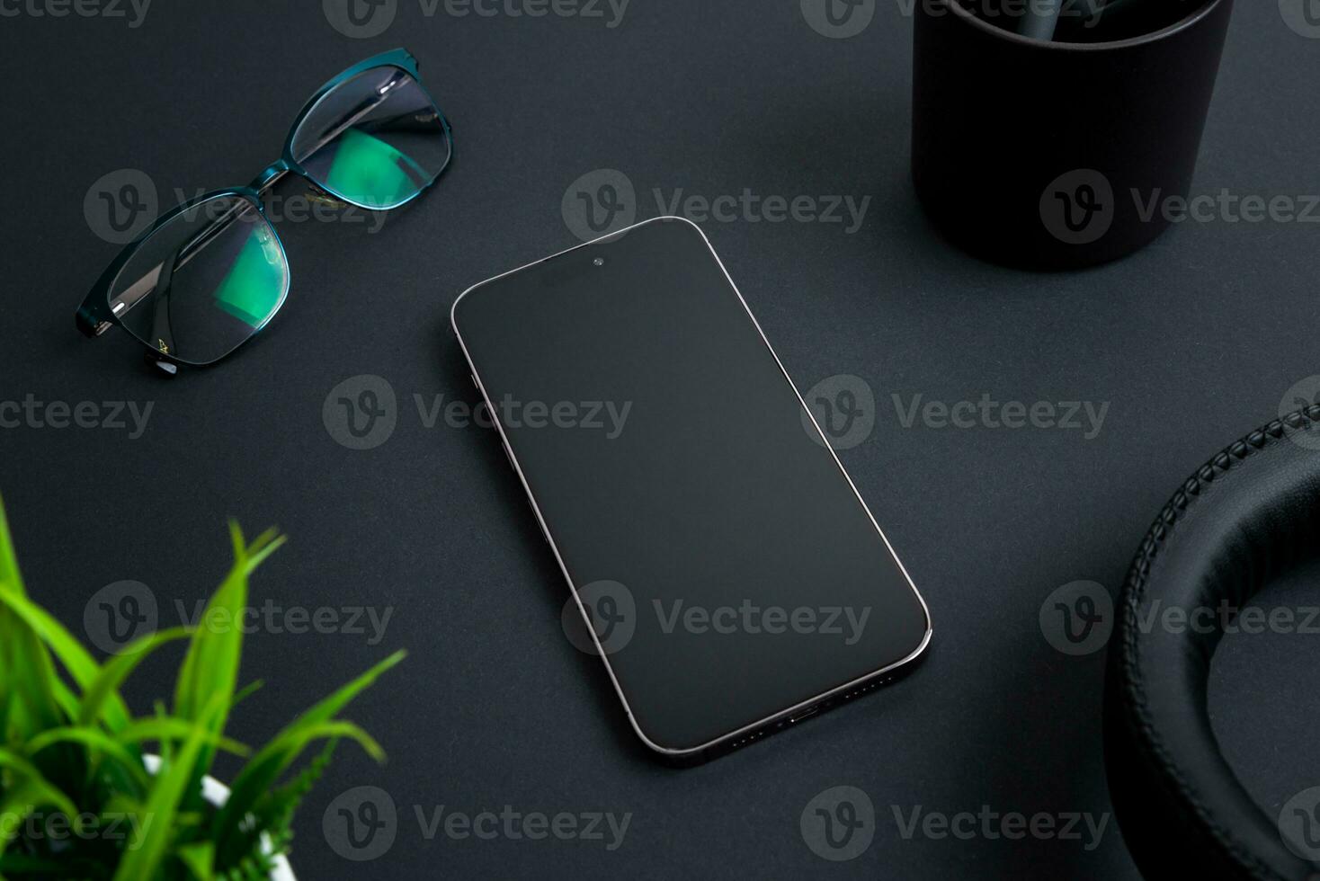 Phone placed on black desk among office items like a headset, plant, glasses, and a coffee mug photo