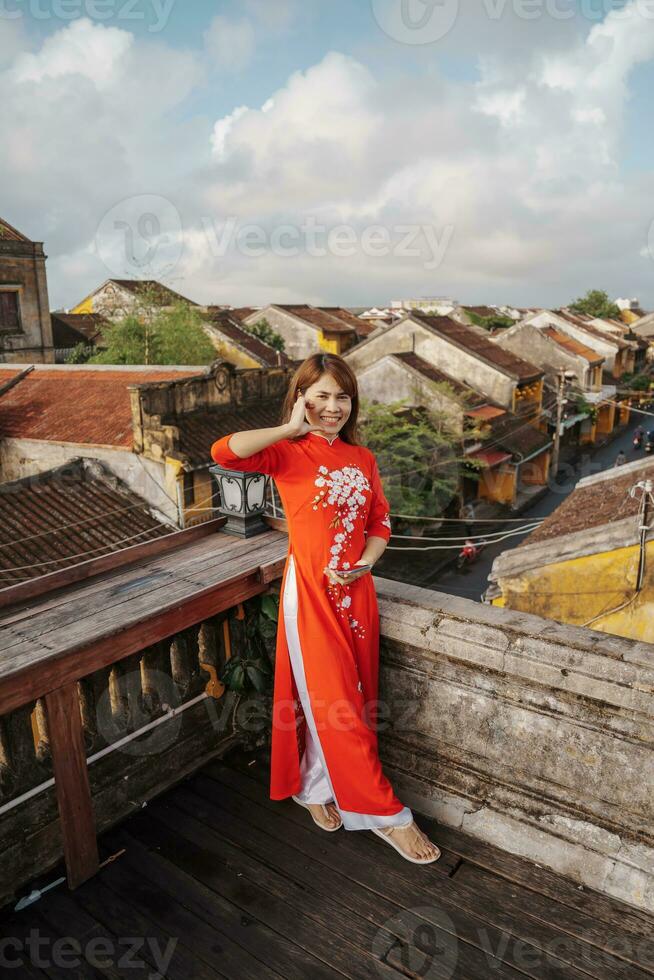 happy woman wearing Ao Dai Vietnamese dress, traveler sightseeing view at rooftop at Hoi An ancient town in Vietnam. landmark and popular for tourist attractions. Vietnam and Southeast travel concept photo