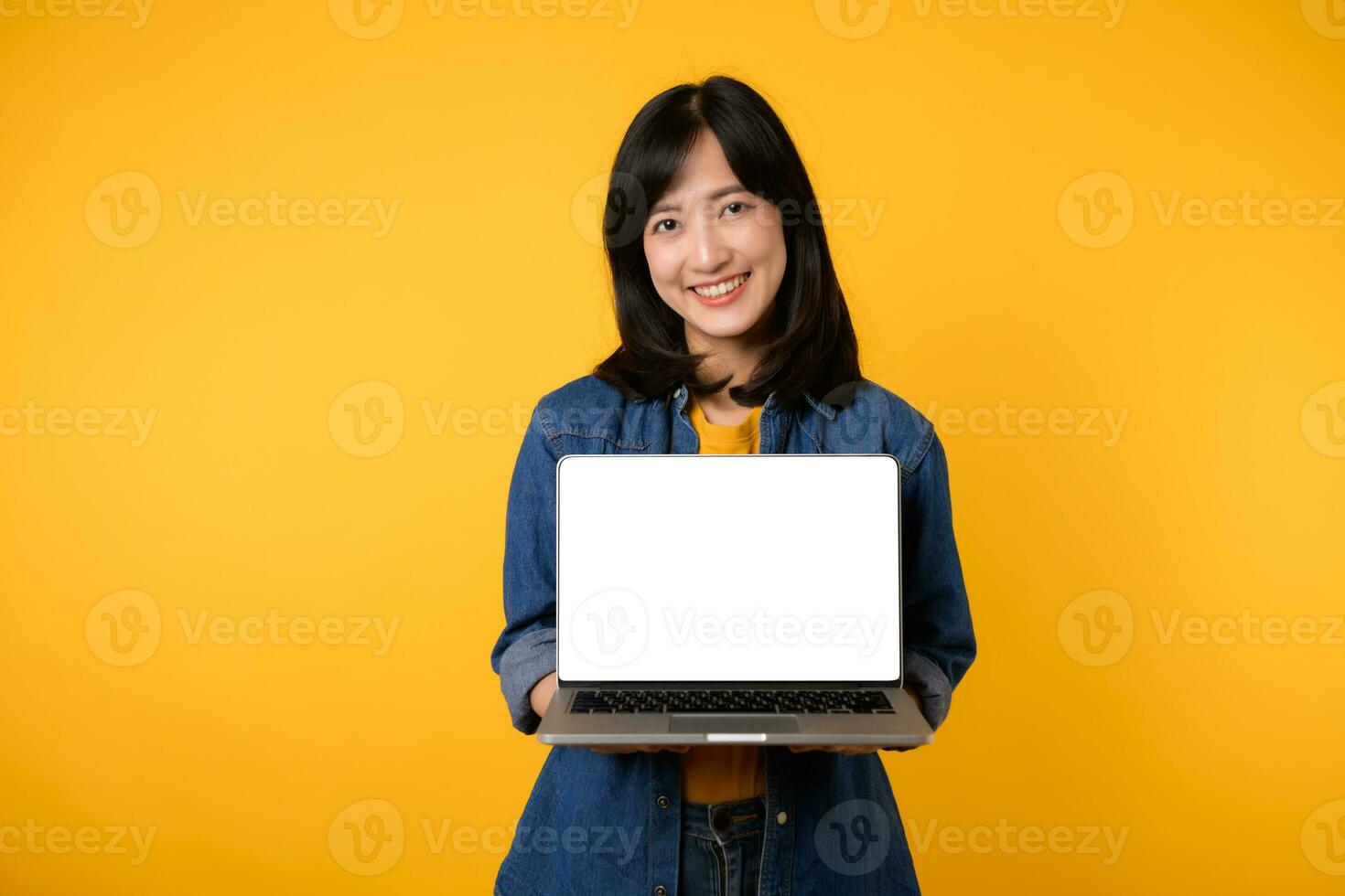 portrait young happy woman wearing yellow t-shirt and denim shirt holding laptop and point finger to screen isolated on yellow studio background. business technology application communication concept. photo