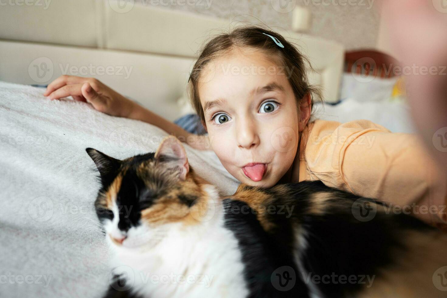 Little girl taking selfie with cat on bed at home. Portrait of adorable child show tongue with pet. photo