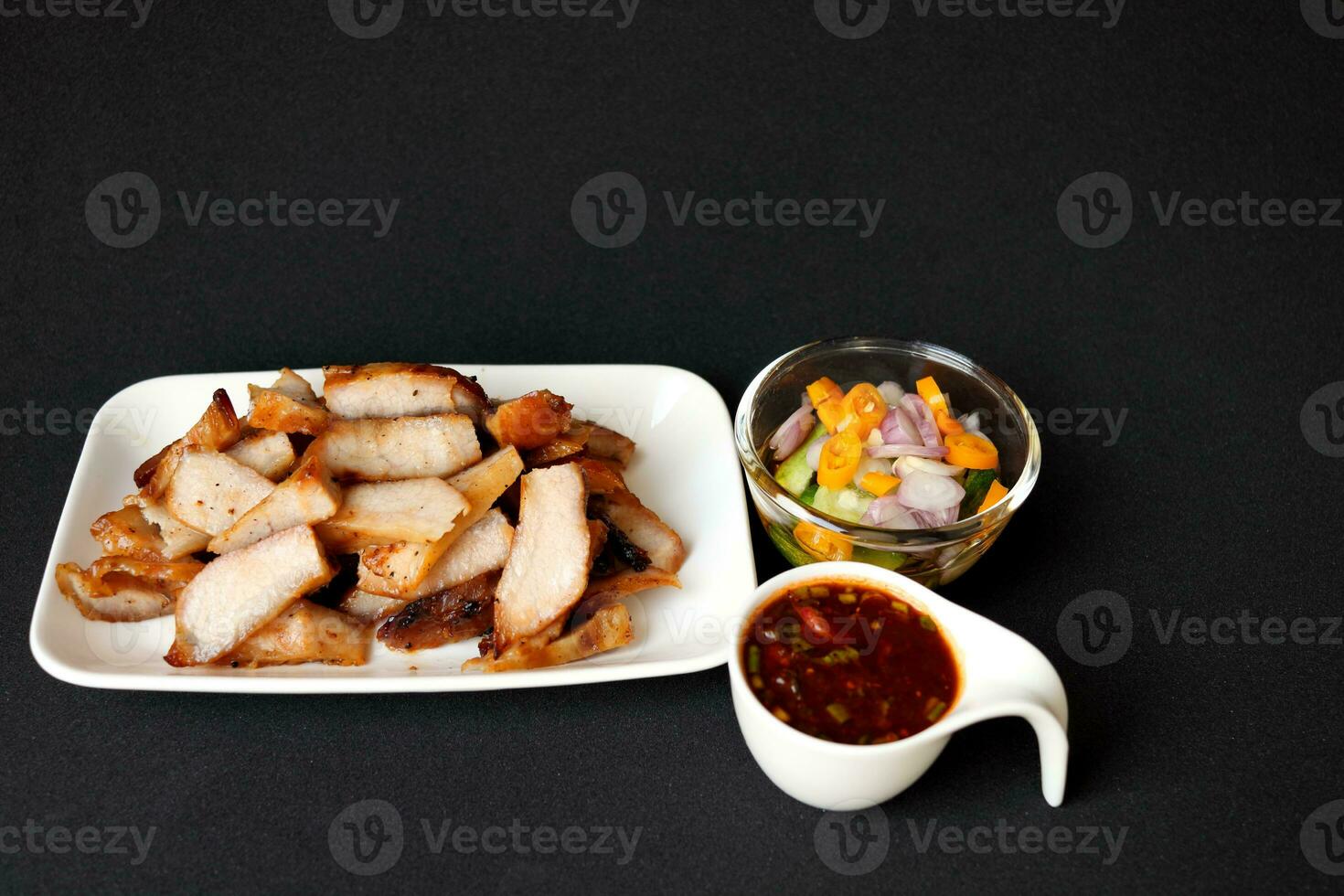 Grilled pork shoulder slice in white rectangle dish with two spicy Thai style sauce in white cup glass cup on black background. photo