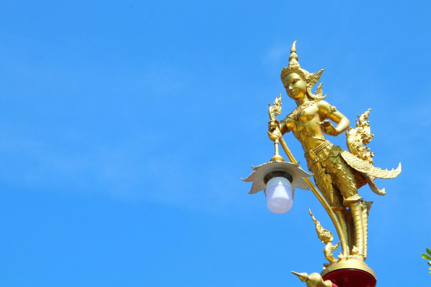 Golden color painted Kinnaree statue holding lamp at the top of pillar and bright blue sky background. Kinnaree is female bird with human head in ancient Thai tale. photo