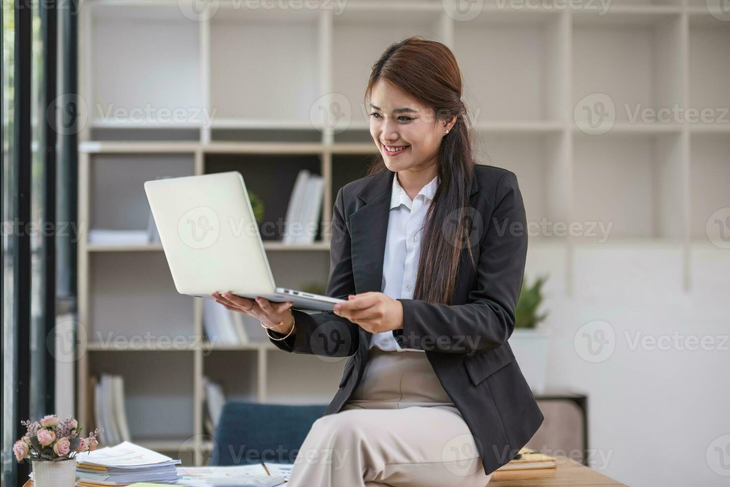 retrato, profesional y confidente milenario asiático mujer de negocios o hembra ejecutivo gerente en formal traje y los anteojos, de pie, propensión en mesa, participación ordenador portátil y utilizando ordenador portátil computadora. foto