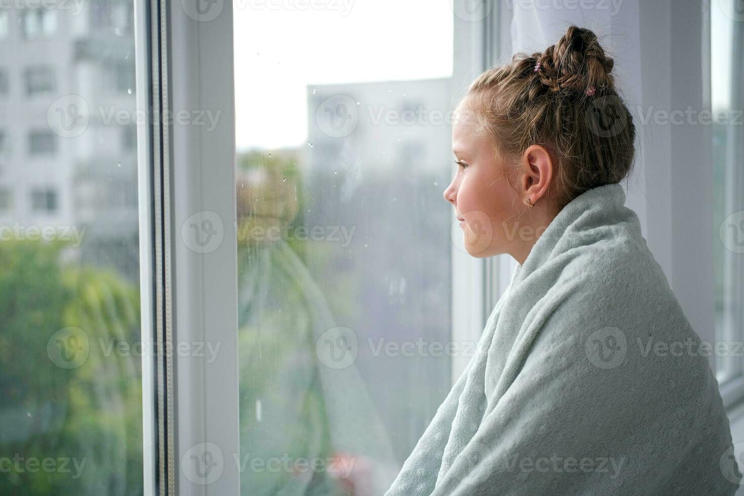 The girl looks out the window at the rain. The child is sad near the window because of the bad weather. photo