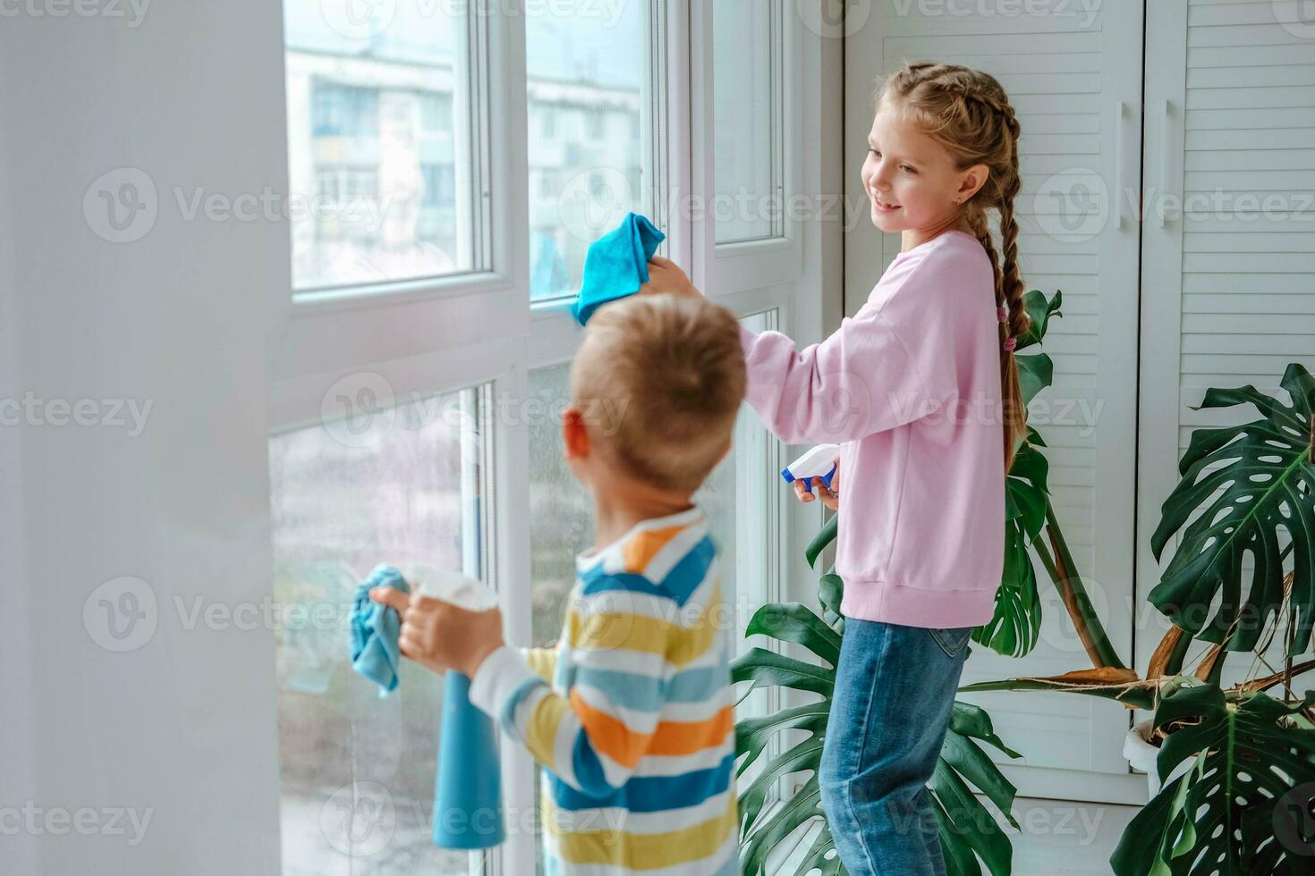 pequeño chico y niña asperja agua desde un botella en el ventana y toallitas eso apagado con un trapo. el niños ayuda con limpieza el casa. limpiar el ventana. hermano y hermana ocupado limpieza. foto