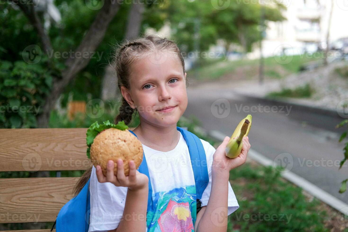 A schoolgirl girl sits on a bench and cannot choose what to eat. Choice of healthy or unhealthy food. Child chooses between burger and banana photo