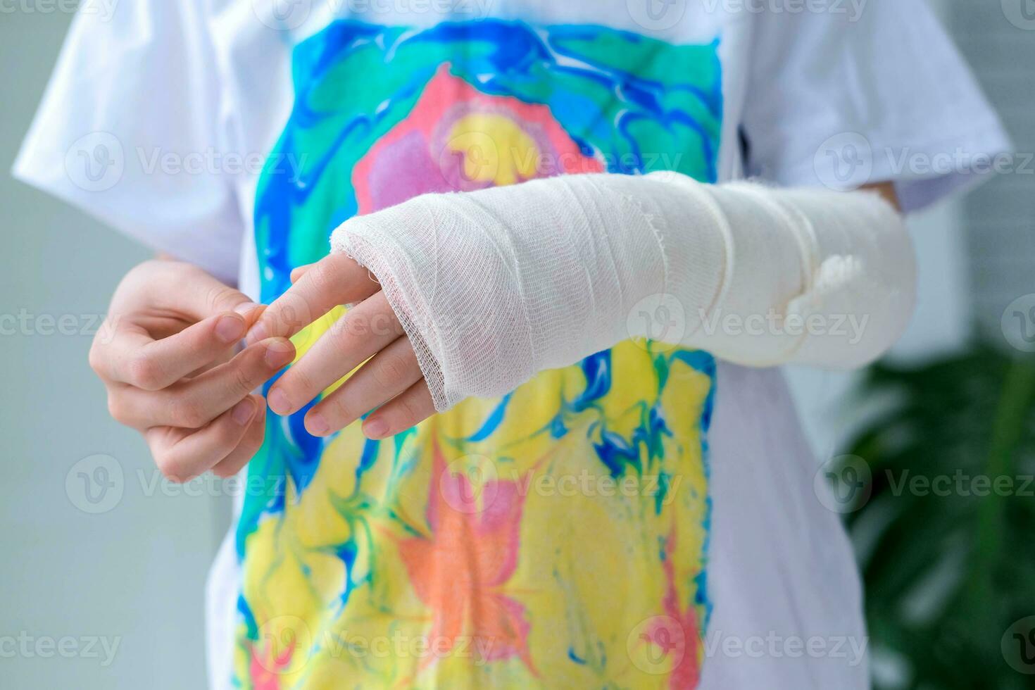 Child doing massage on fingers of broken hand wrapped in white plaster bandage.  Concept of healthcare. The girl holds a broken arm against the background of a multi-colored t-shirt photo