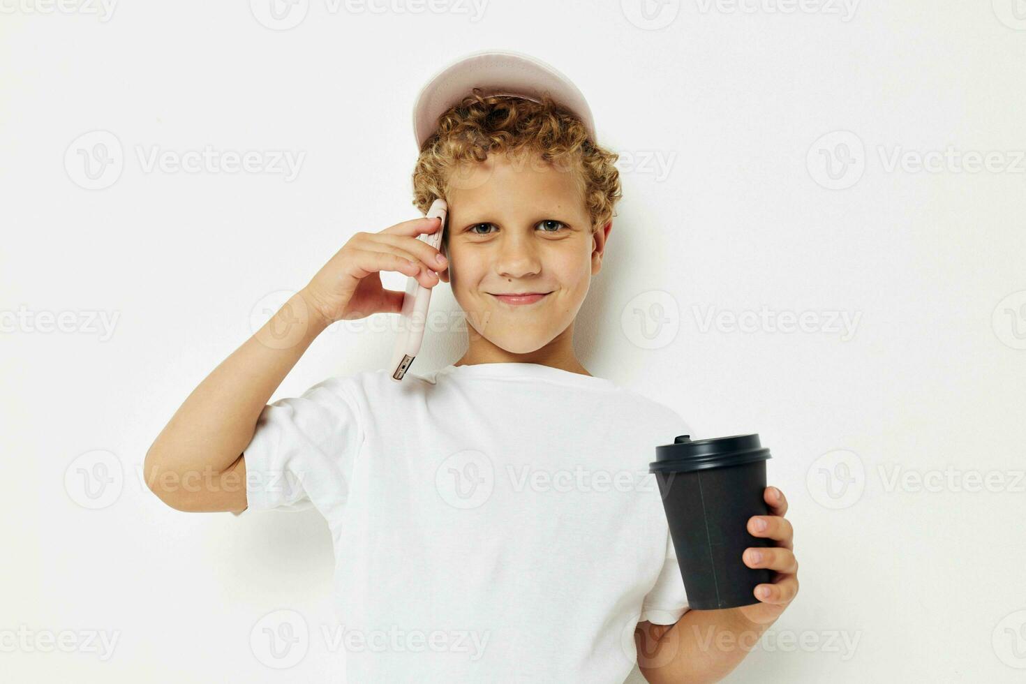 Photo portrait curly little boy in a white t-shirt cap with a phone in a glass with a drink light background unaltered