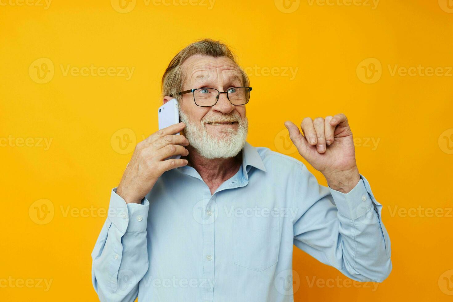 Photo of retired old man in a blue shirt and glasses talking on the phone yellow background