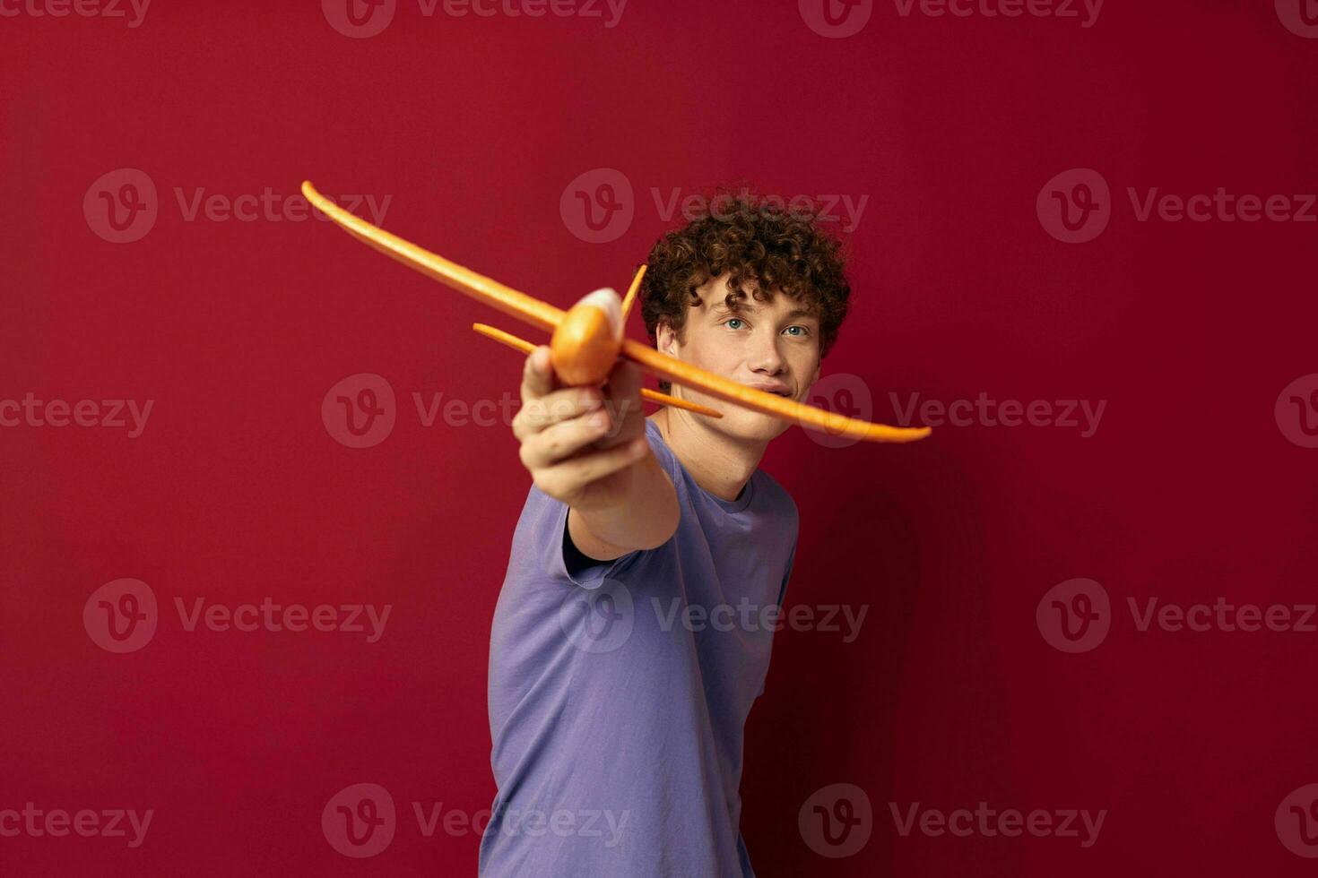 A young man with a toy plane in a purple t-shirt red background unaltered photo